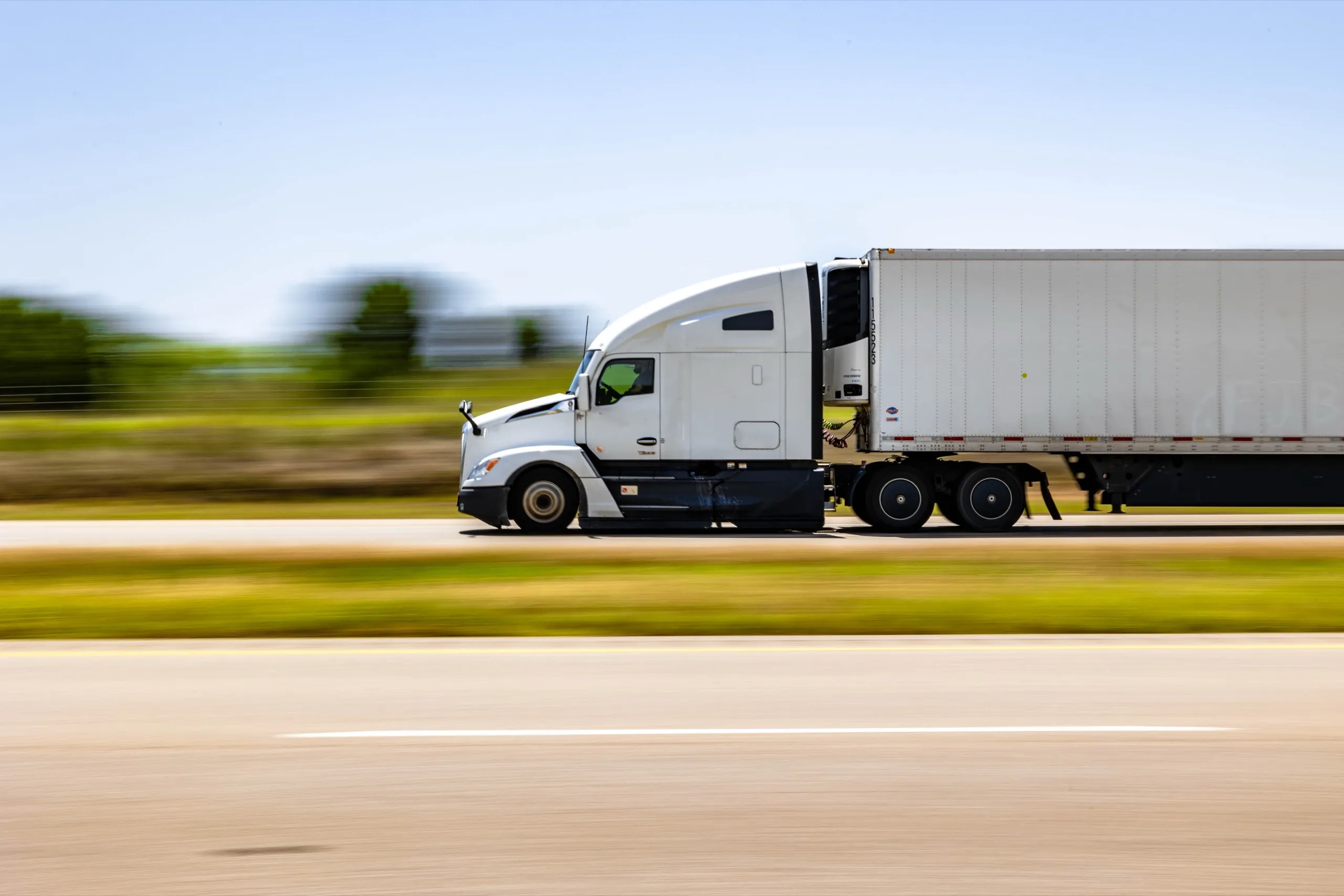 White semi truck driving fast along a rural highway with blurred green trees in the background.
