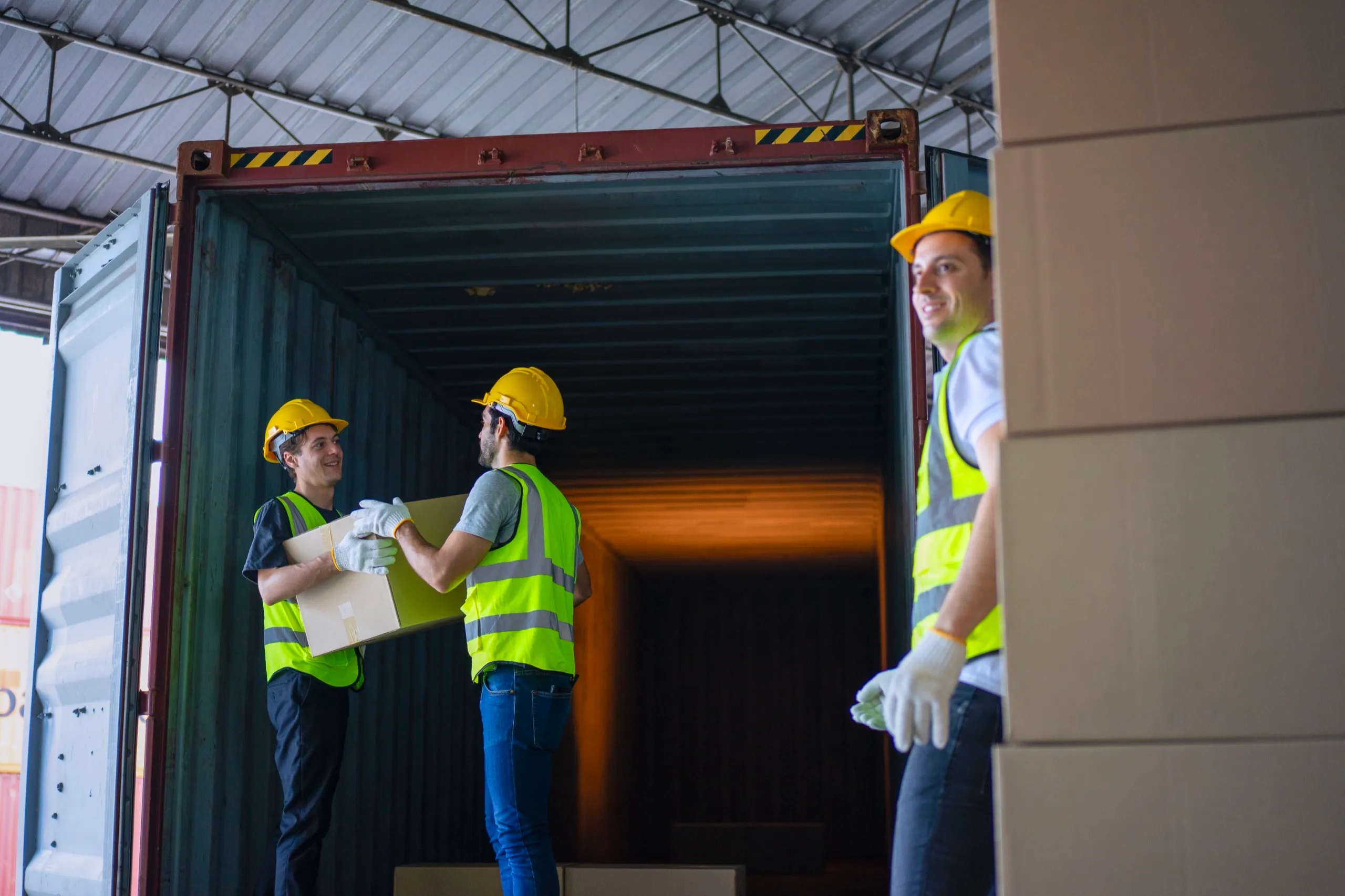 Three warehouse workers wearing yellow hard hats and reflective vests, two handing a cardboard box inside a shipping container and one standing beside stacked boxes.