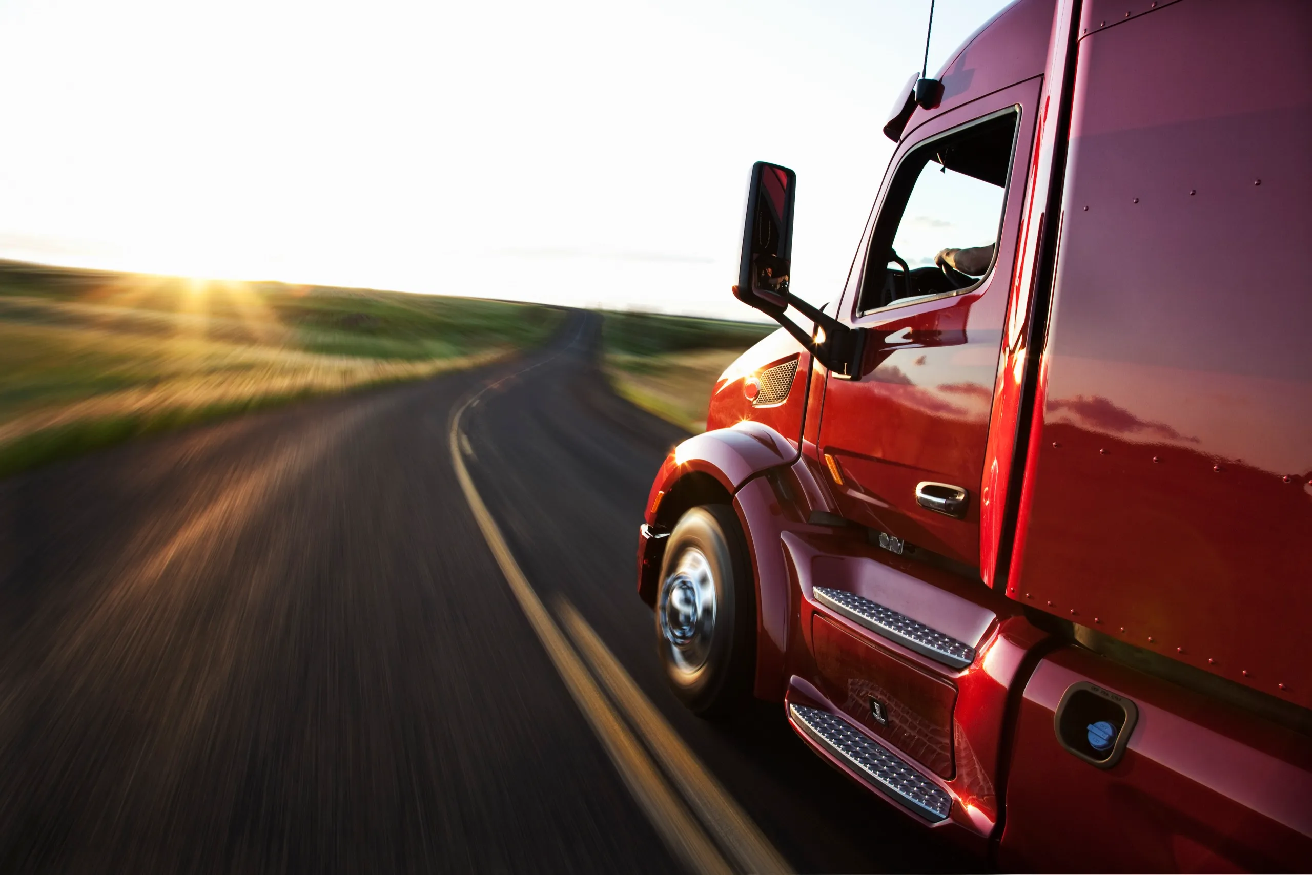 Close-up side view of a red commercial truck driving on a winding road during sunset.