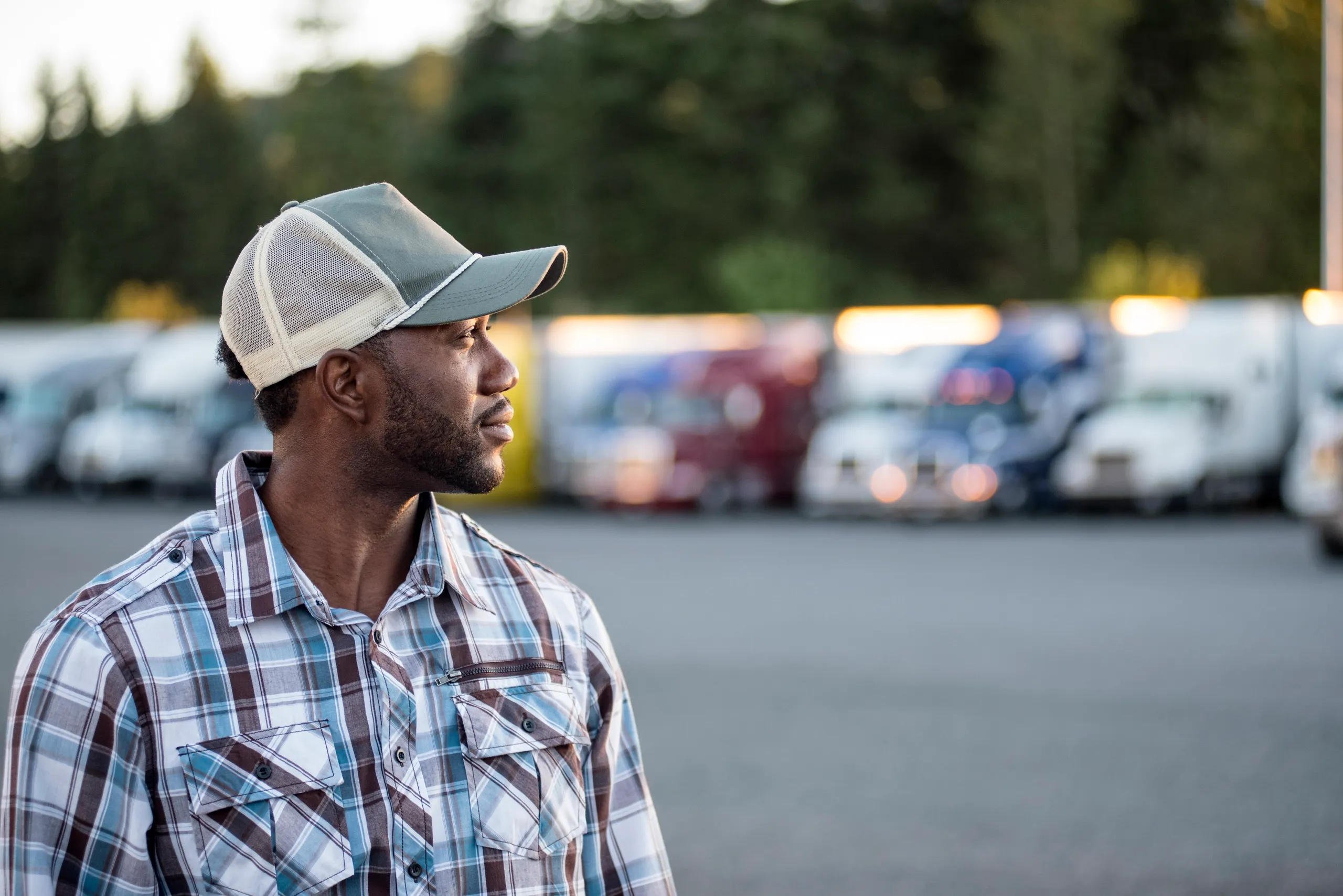 Man in a plaid shirt and baseball cap standing outdoors with parked trucks blurred in the background.