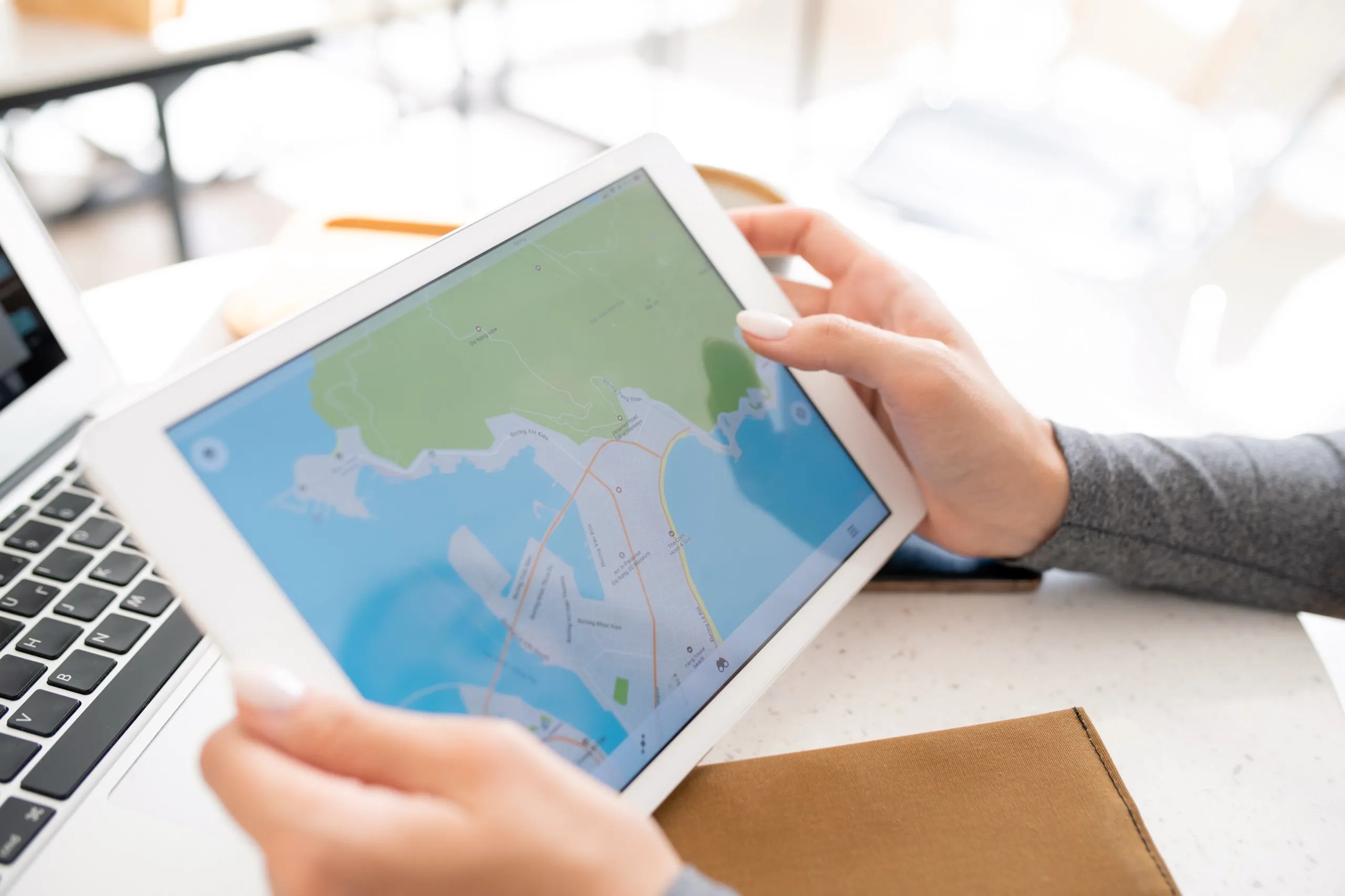 Hands of a person holding a tablet displaying a map, next to a laptop and a brown notebook on a desk.