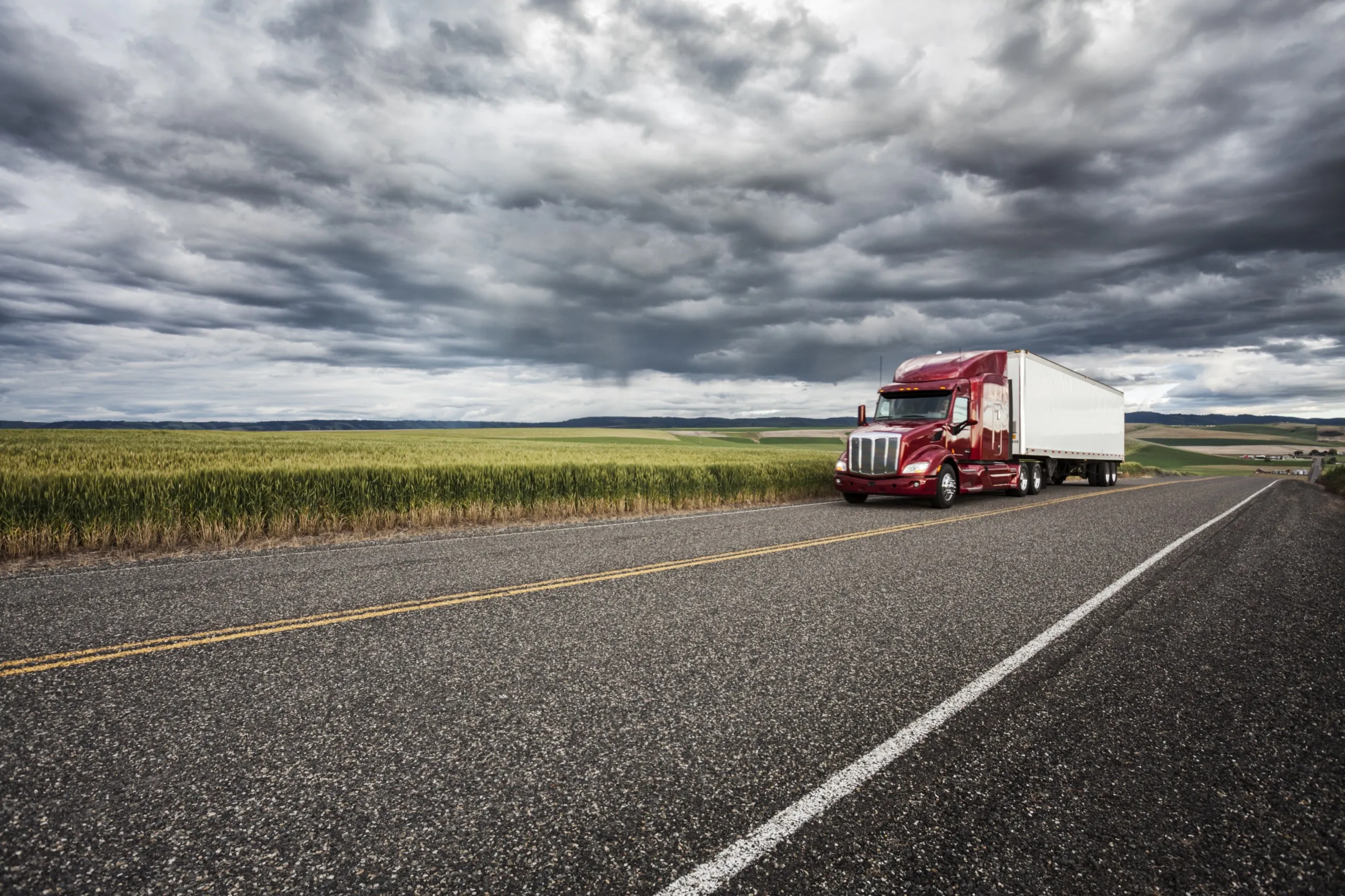 Red semi-truck driving on a rural highway beside green wheat fields under a cloudy sky.