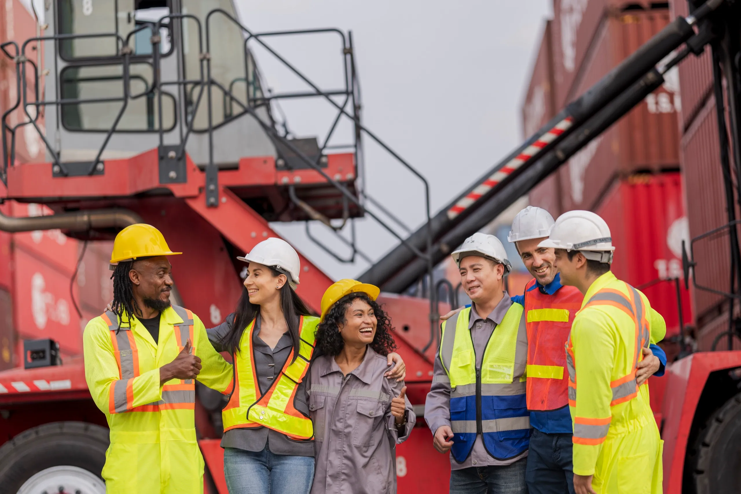 Group of six diverse construction workers wearing safety helmets and high-visibility vests, smiling and interacting near red industrial machinery and shipping containers.