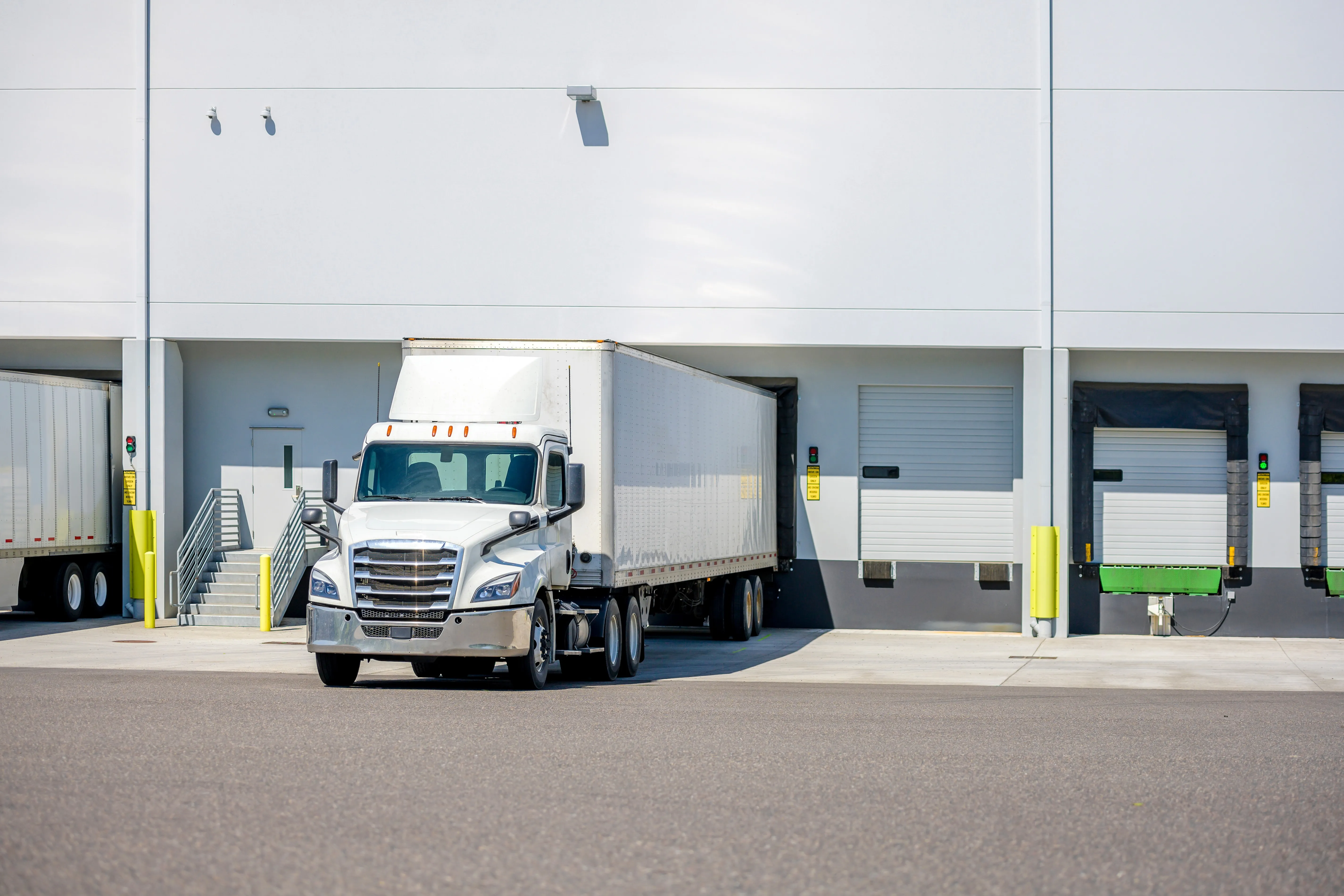 White semi-truck backed into a loading dock at a warehouse with multiple closed dock doors.