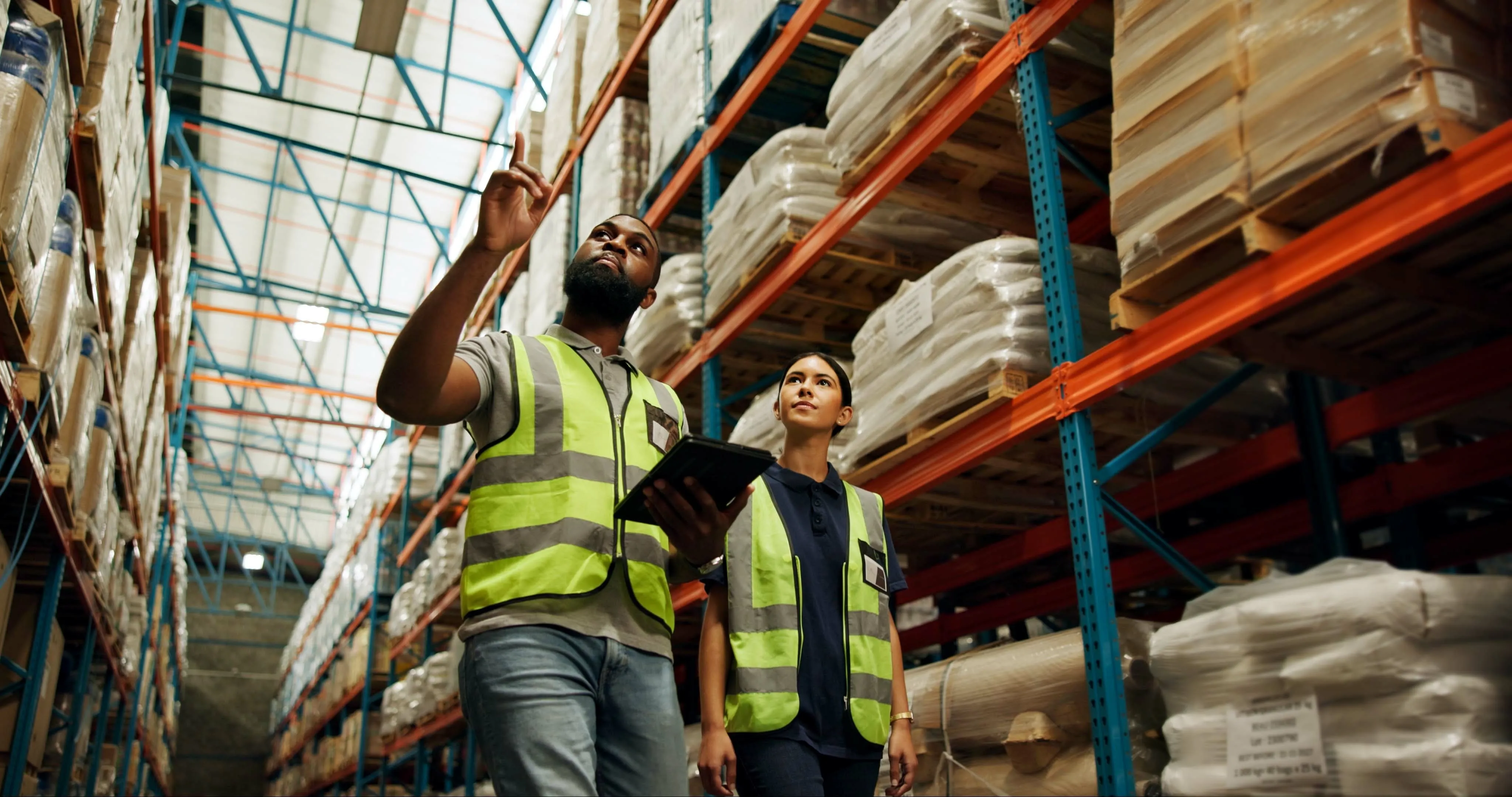 Two warehouse workers wearing reflective safety vests inspecting and discussing inventory on tall shelves, one holding a tablet.
