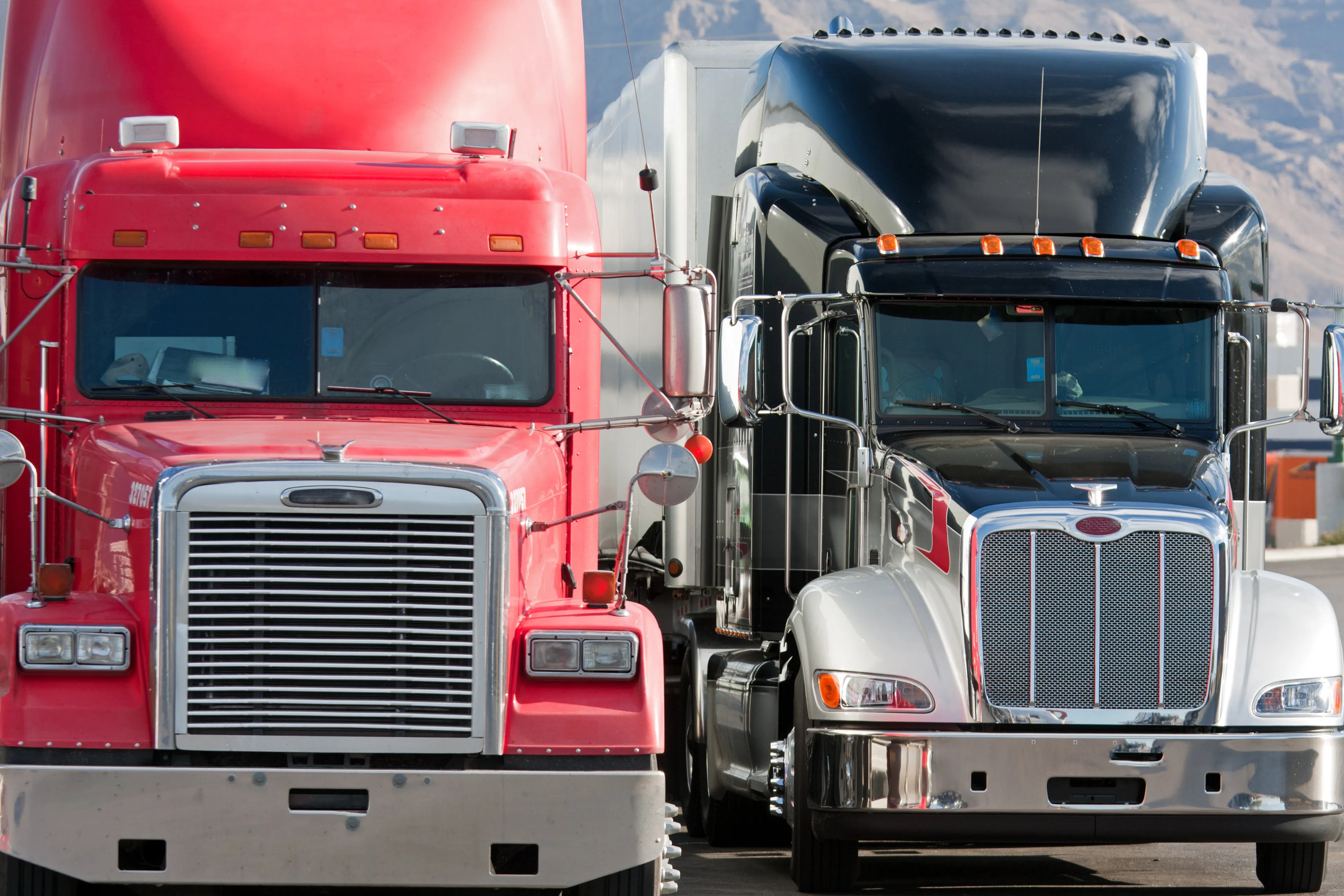 Close-up of a red semi-truck and a black and white semi-truck parked side by side against a mountainous background.