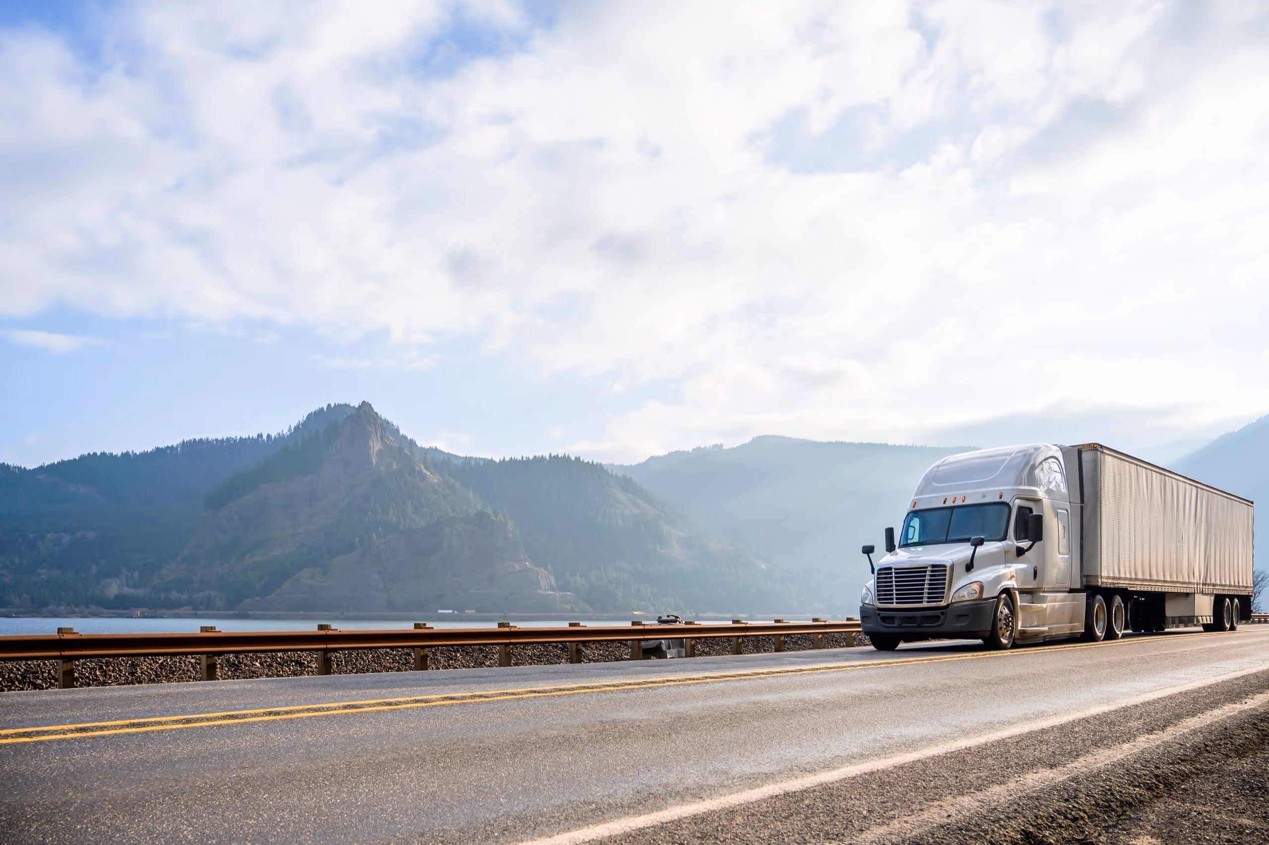 Semi-truck driving on a highway beside a lake with forested mountains under a partly cloudy sky.