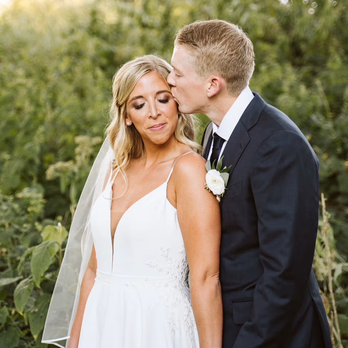 Groom in black suit kissing the bride's temple as she smiles with eyes closed outdoors.