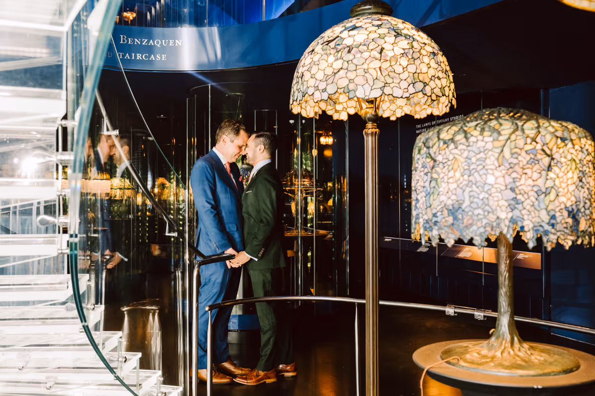 Two men in suits holding hands and touching foreheads inside a modern glass staircase area with Tiffany-style lamps.