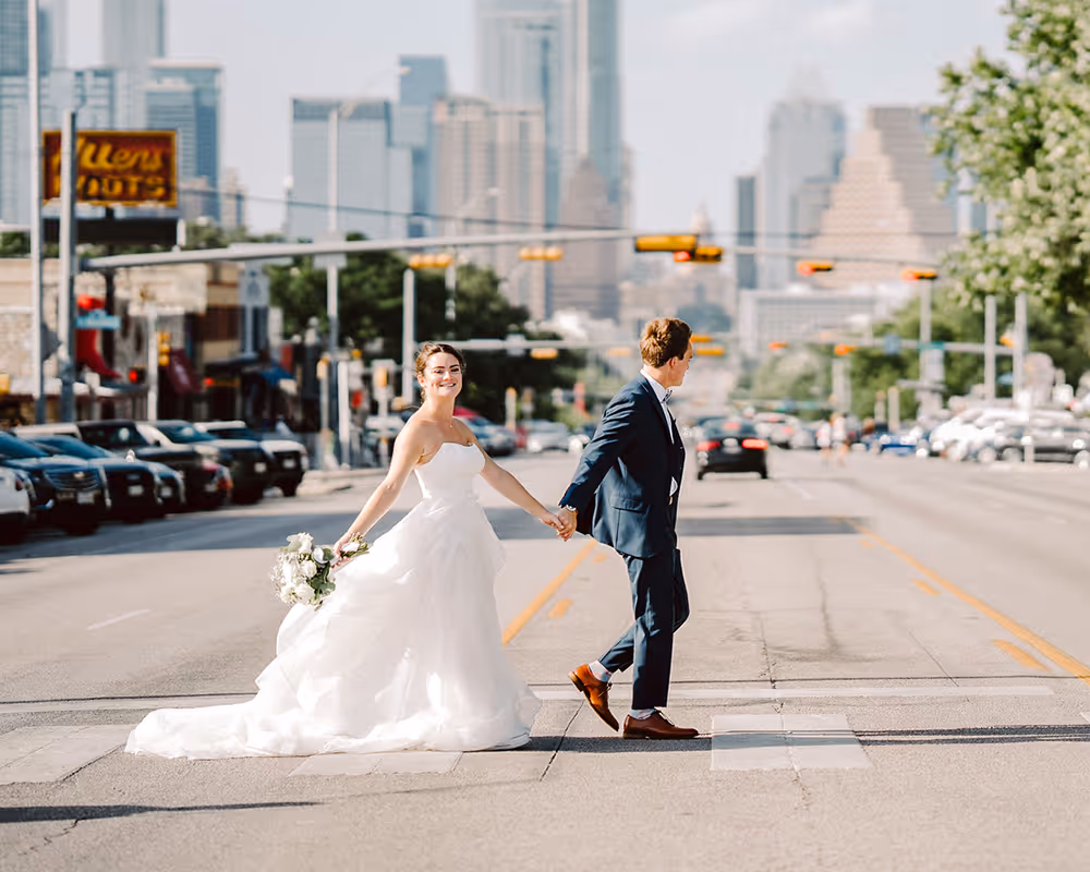 Bride in a white wedding dress holding a bouquet and smiling, holding hands with groom in a navy suit, walking across a city street with tall buildings in the background.