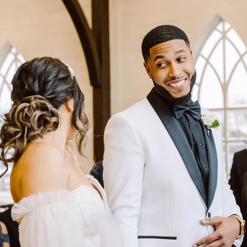 Groom in a white tuxedo with black lapels smiling at bride in a white wedding dress with styled hair inside a building with arched windows.