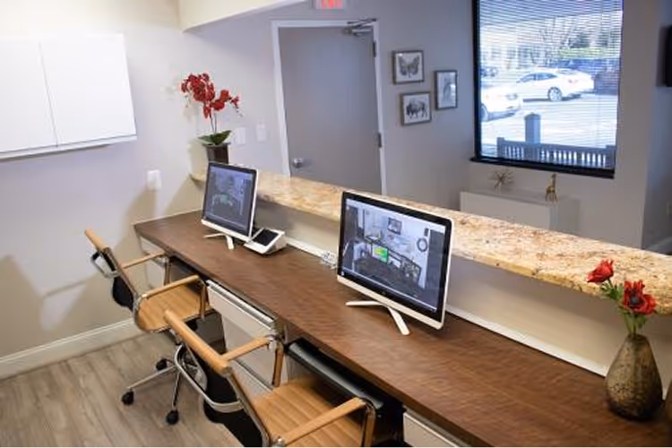 Modern office workspace with two wooden chairs, computers on a dark wooden desk, a granite countertop, and a window showing parked cars outside.