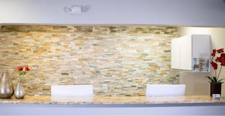 Reception desk with beige stone wall backdrop, two white chairs, silver vases, and red flowers.