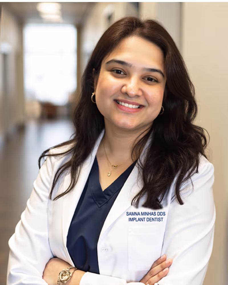 Smiling female implant dentist in white coat and navy scrubs standing with arms crossed in a hallway.