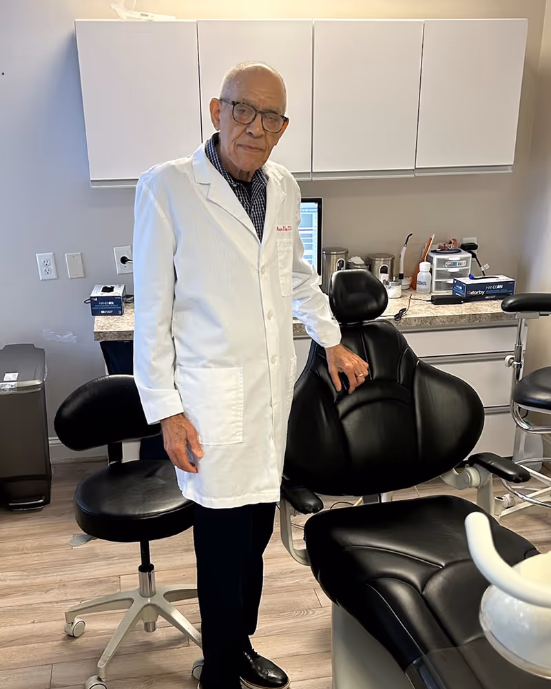 Elderly man in glasses and white lab coat standing next to a black leather dental chair in a dental office.