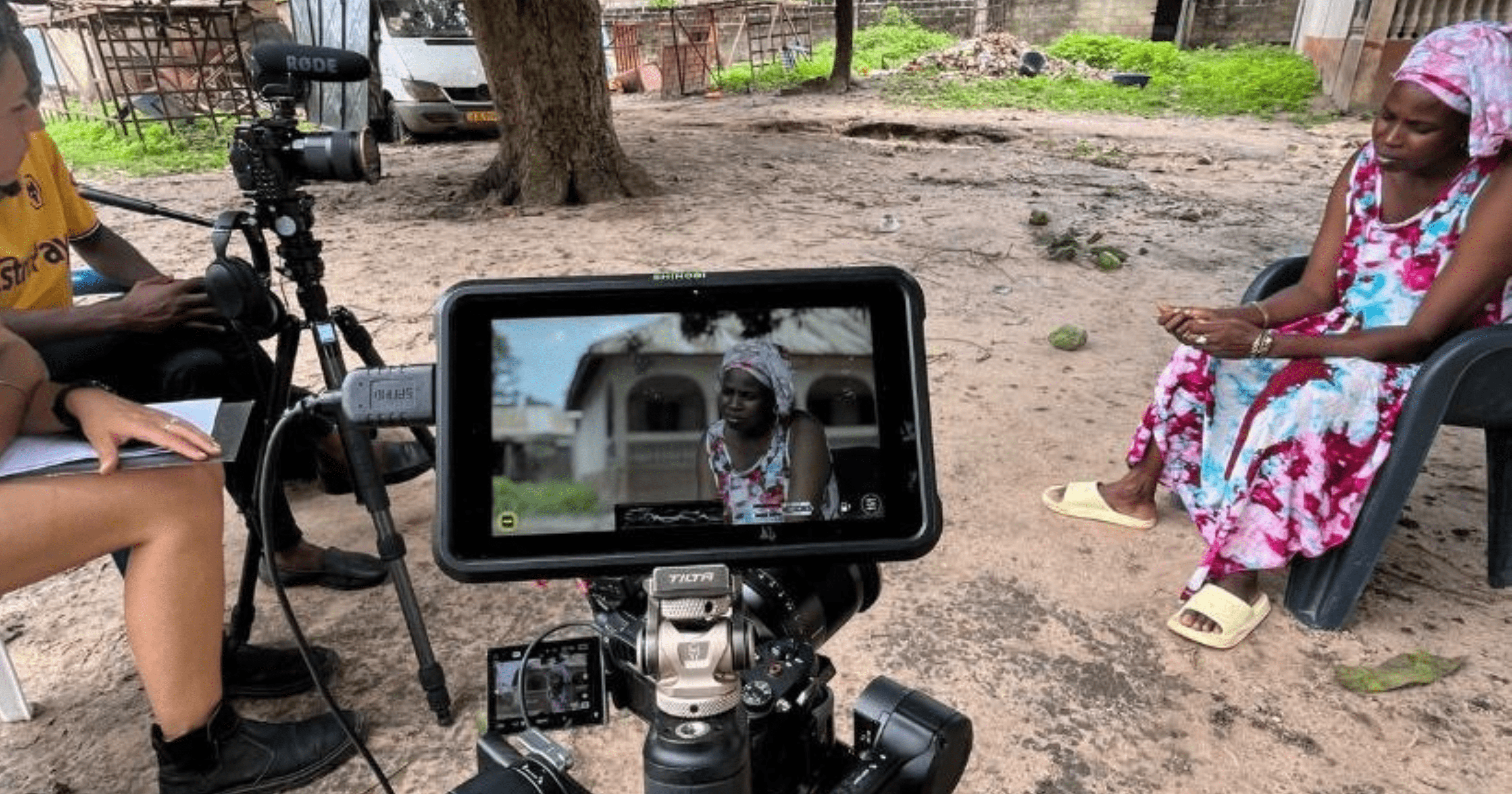 A woman in a colorful dress and headscarf sits on a plastic chair outdoors, while a camera on a tripod films her and displays her image on a monitor.