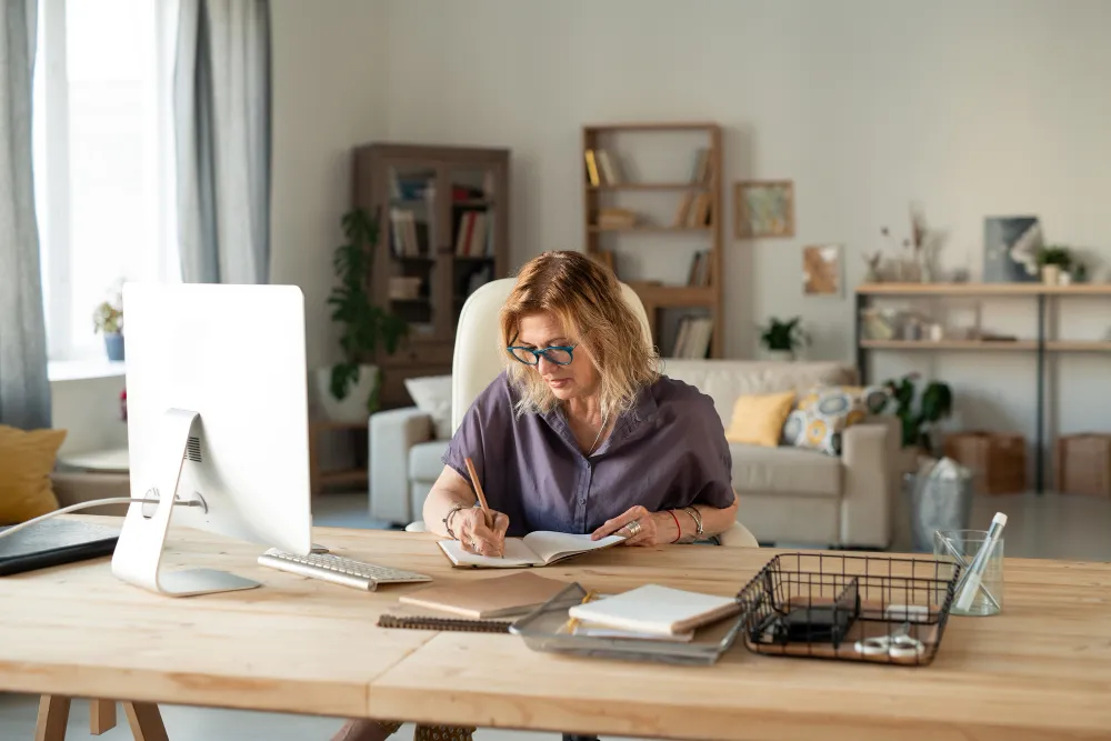 Woman writing notes at a desk with desktop computer in a bright home office living space