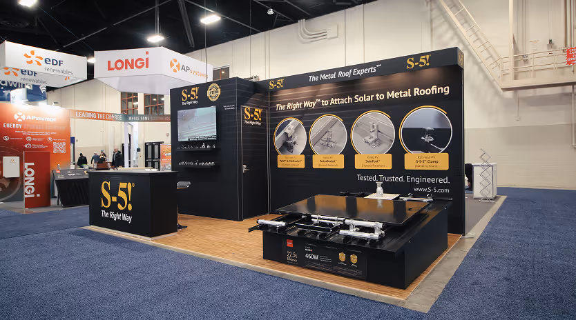 Trade show booth for S-5! showcasing metal roof solar attachment products with display screens and informational graphics.