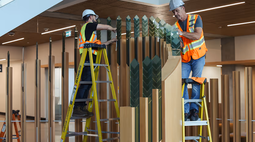 Two construction workers in safety vests and helmets assembling or installing a large decorative wooden and green panel inside a modern building.