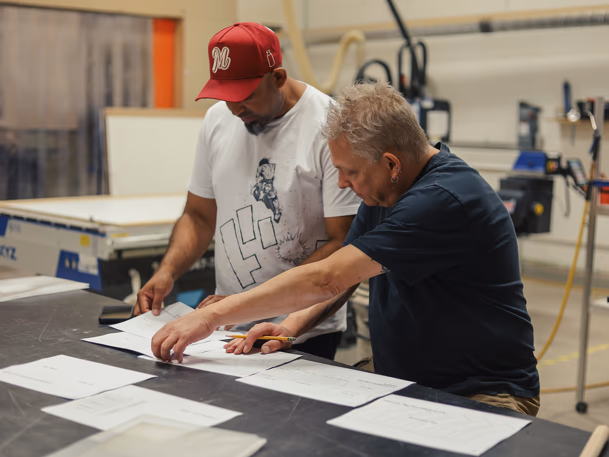 Two men in a workshop reviewing and organizing multiple printed documents laid out on a table.