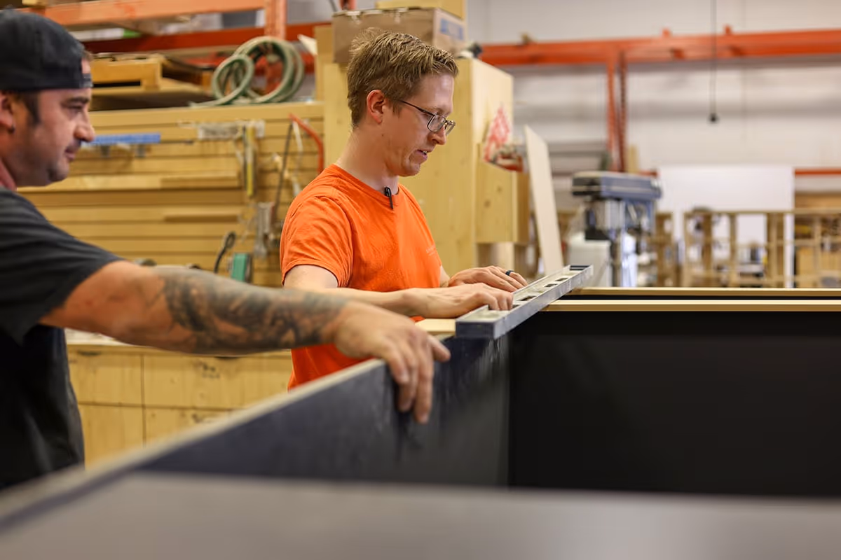 Two men working in a woodshop using a level tool to check a large black surface.