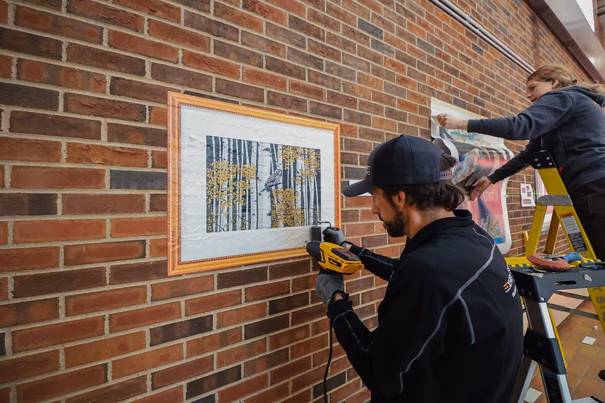 A man using a heat press machine on a framed picture of tree trunks with yellow leaves on a brick wall, while a woman on a ladder applies a large printed graphic.