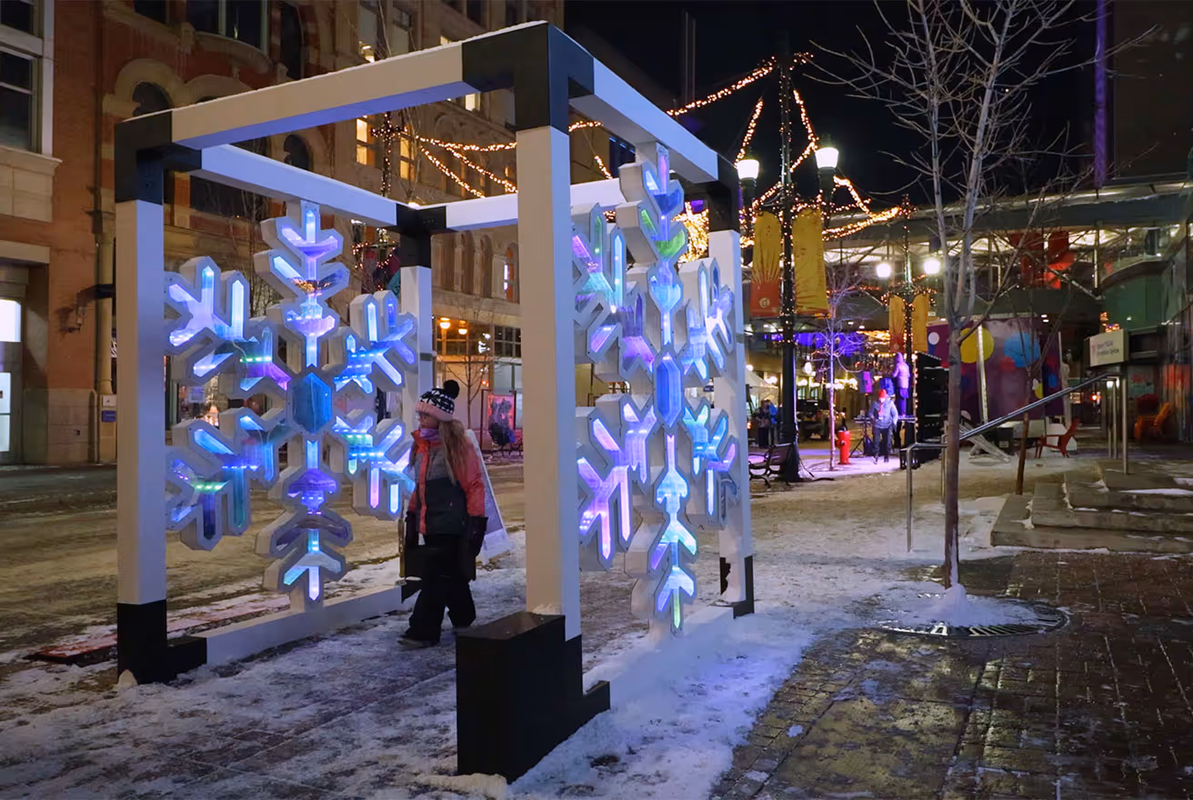Person walking through a large illuminated snowflake installation on a snowy city street at night.