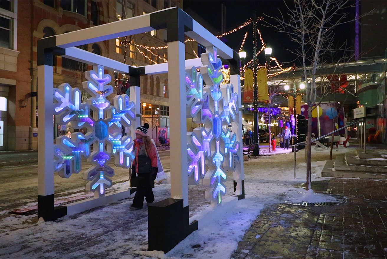 Person walking through a large illuminated snowflake installation on a snowy city street at night.