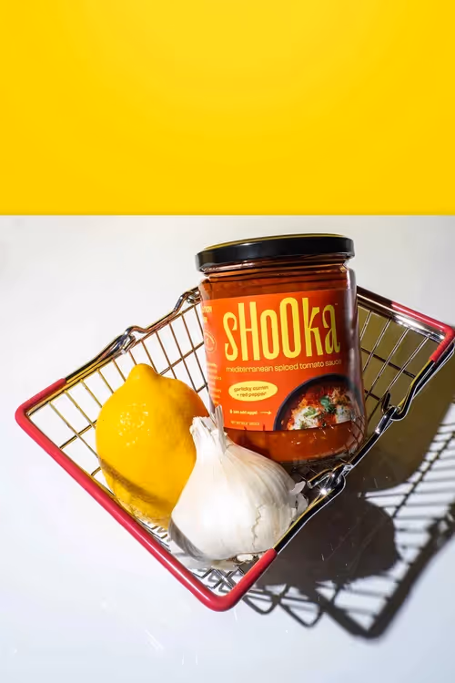 Shooka sauce jar in a wire basket with lemon and garlic on yellow backdrop, food photography by Photos by Lars.