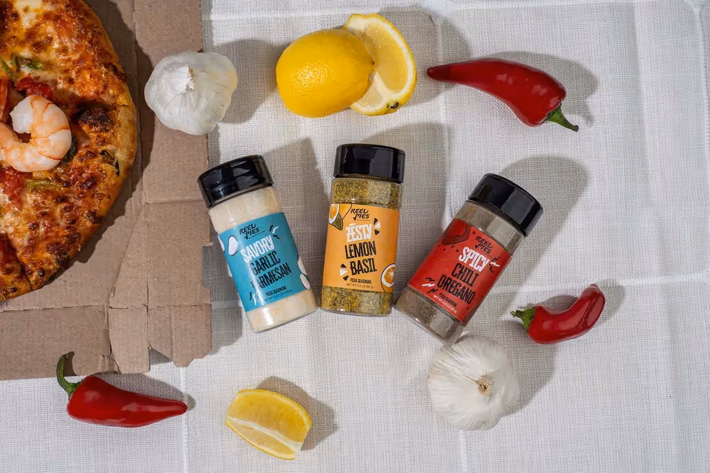 Three spice shakers with chili, lemon, and garlic around a pizza slice on a white cloth, food photography by Photos by Lars.