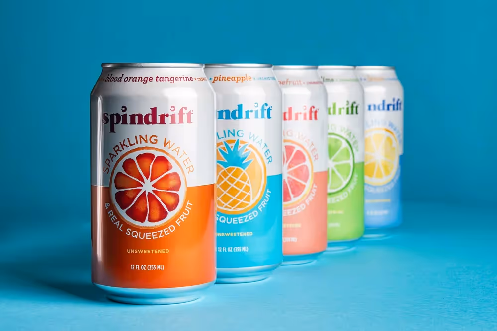 Spindrift sparkling water cans in a row with orange can in front on teal background, product photography by Photos by Lars.