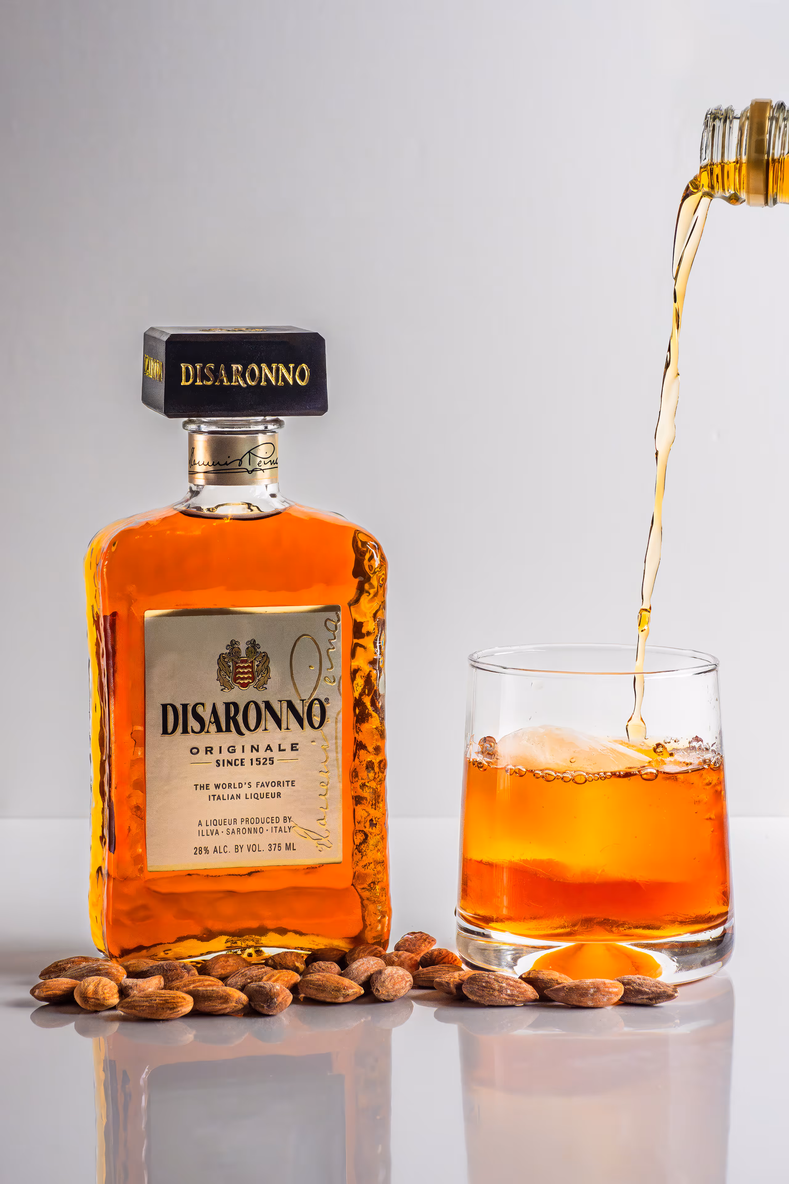 Disaronno bottle pouring into a rocks glass with almonds on white surface, product photography by Photos by Lars.