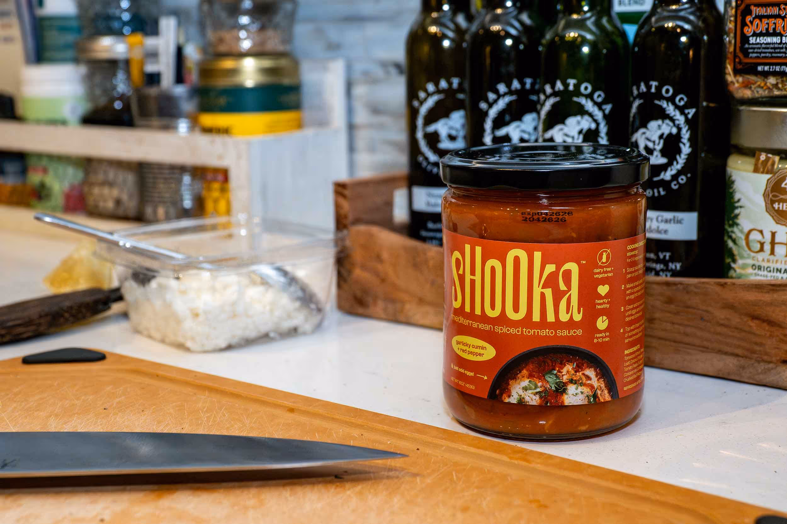 Shooka Mediterranean spiced tomato sauce jar on a kitchen counter beside a knife and sliced cheese, pantry bottles softly blurred behind — Photos by Lars.