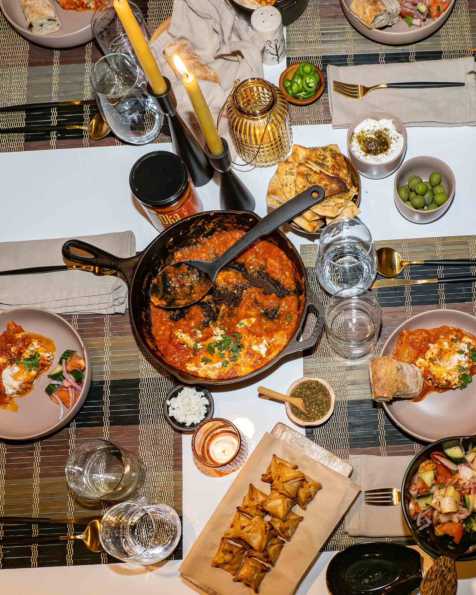 Overhead view of a shared table with a skillet dish being served, surrounded by mezze-style plates and drinks — Photos by Lars.