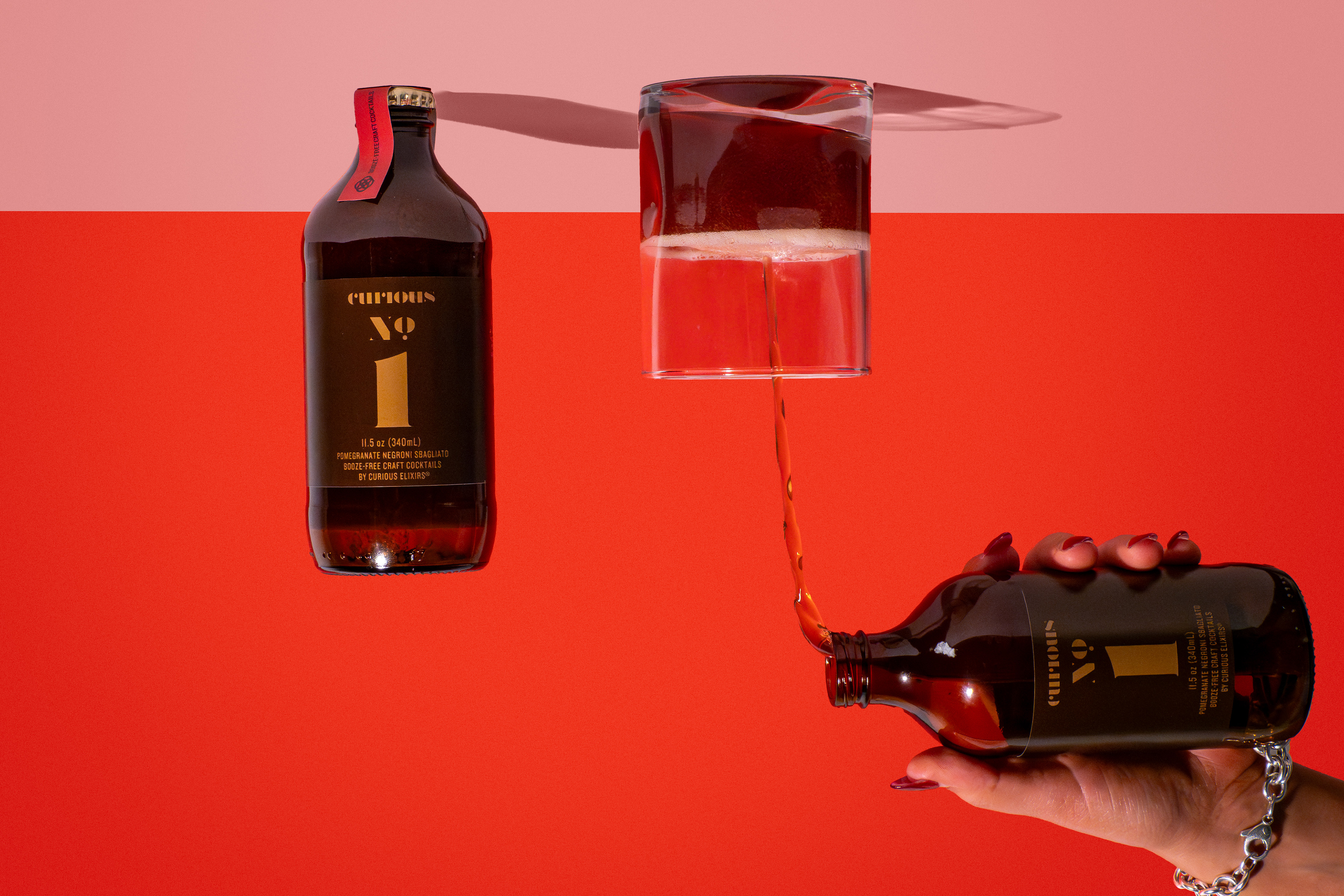 Dark bottle and square cocktail glass suspended over red backdrop with tilted bottle in hand, product photography Photos by Lars.