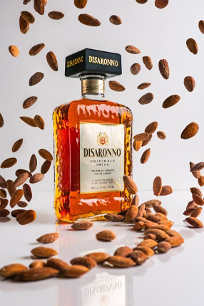 A bottle of Disaronno liqueur stands among scattered almonds against a light background. The glossy liquid shines amber, conveying a sense of luxury and indulgence.