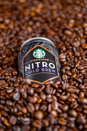 A can of Starbucks Nitro Cold Brew partially buried in a pile of shiny brown coffee beans, conveying a sense of rich aroma and freshness.