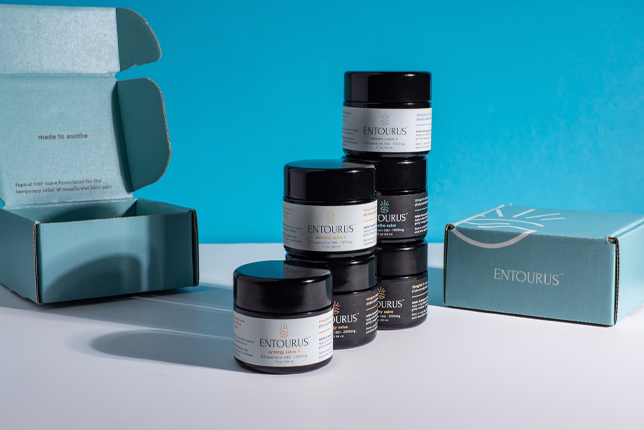 Stack of Entourus product jars on a white surface with a blue background. An open and closed turquoise box sit nearby, conveying a clean, professional tone.