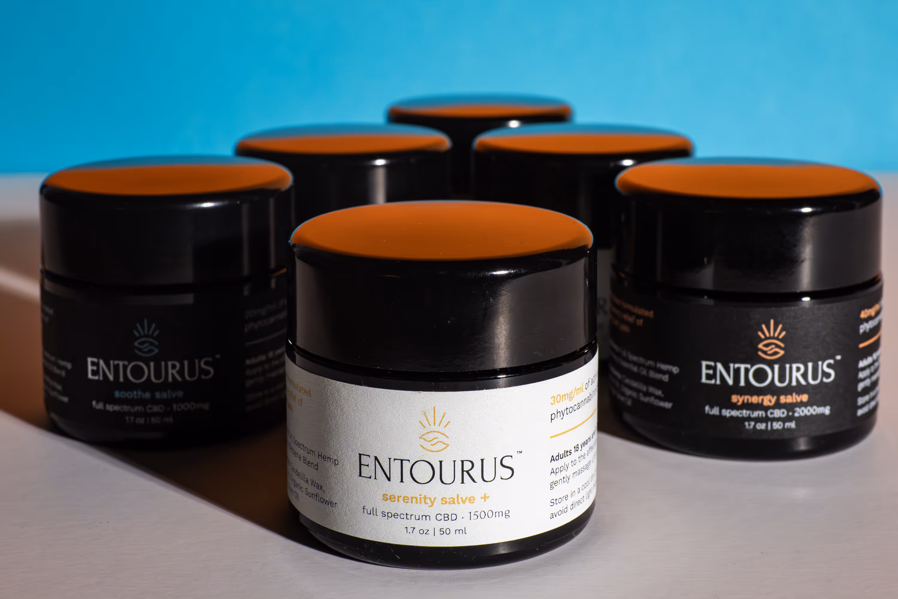 Entourus Serenity Salve+ jar in front of black salve jars with orange lids on blue backdrop, product photography, Photos by Lars.
