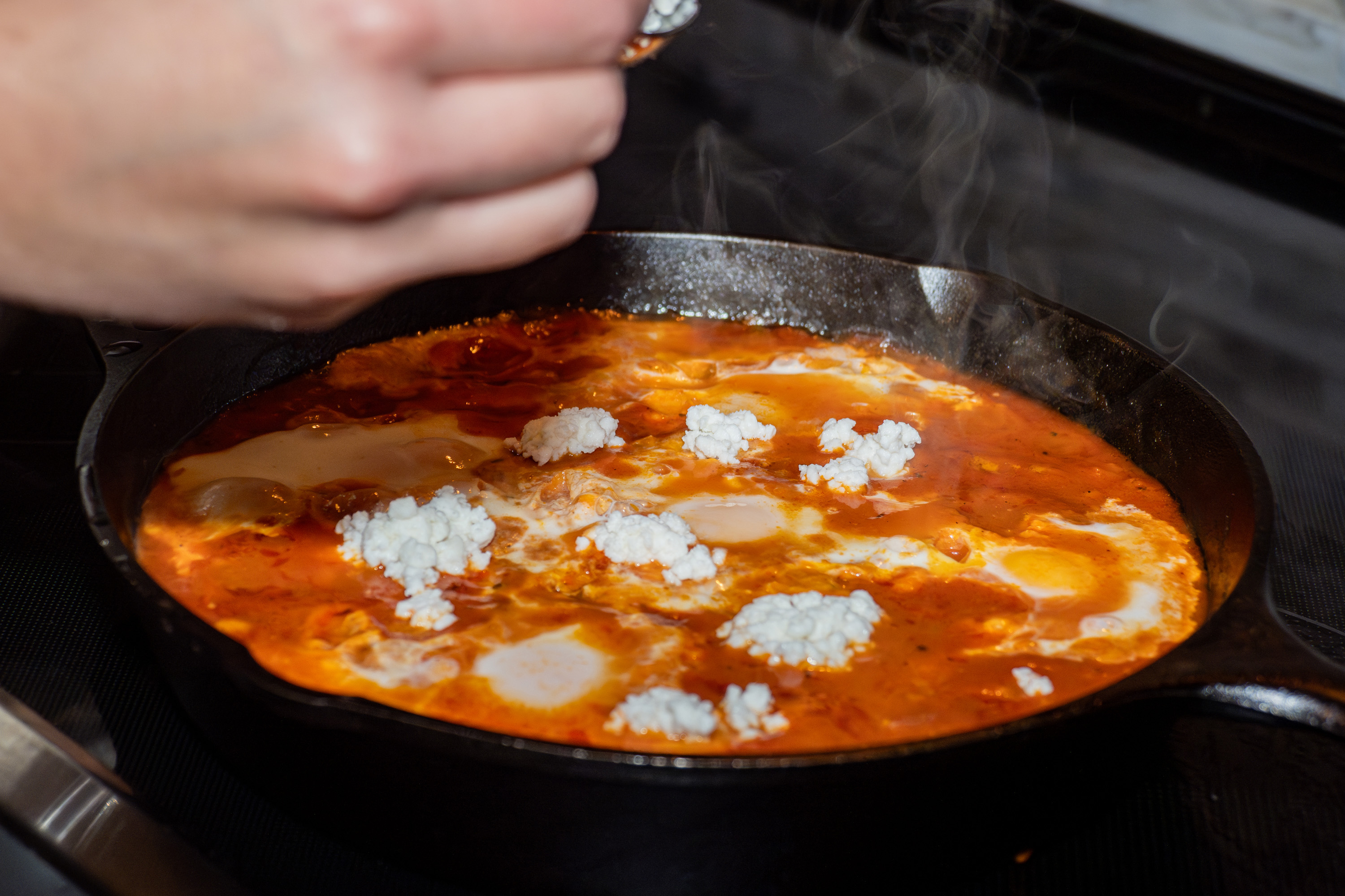 Cast-iron skillet with Shooka sauce, eggs, and white cheese simmering with steam, food photography, Photos by Lars.