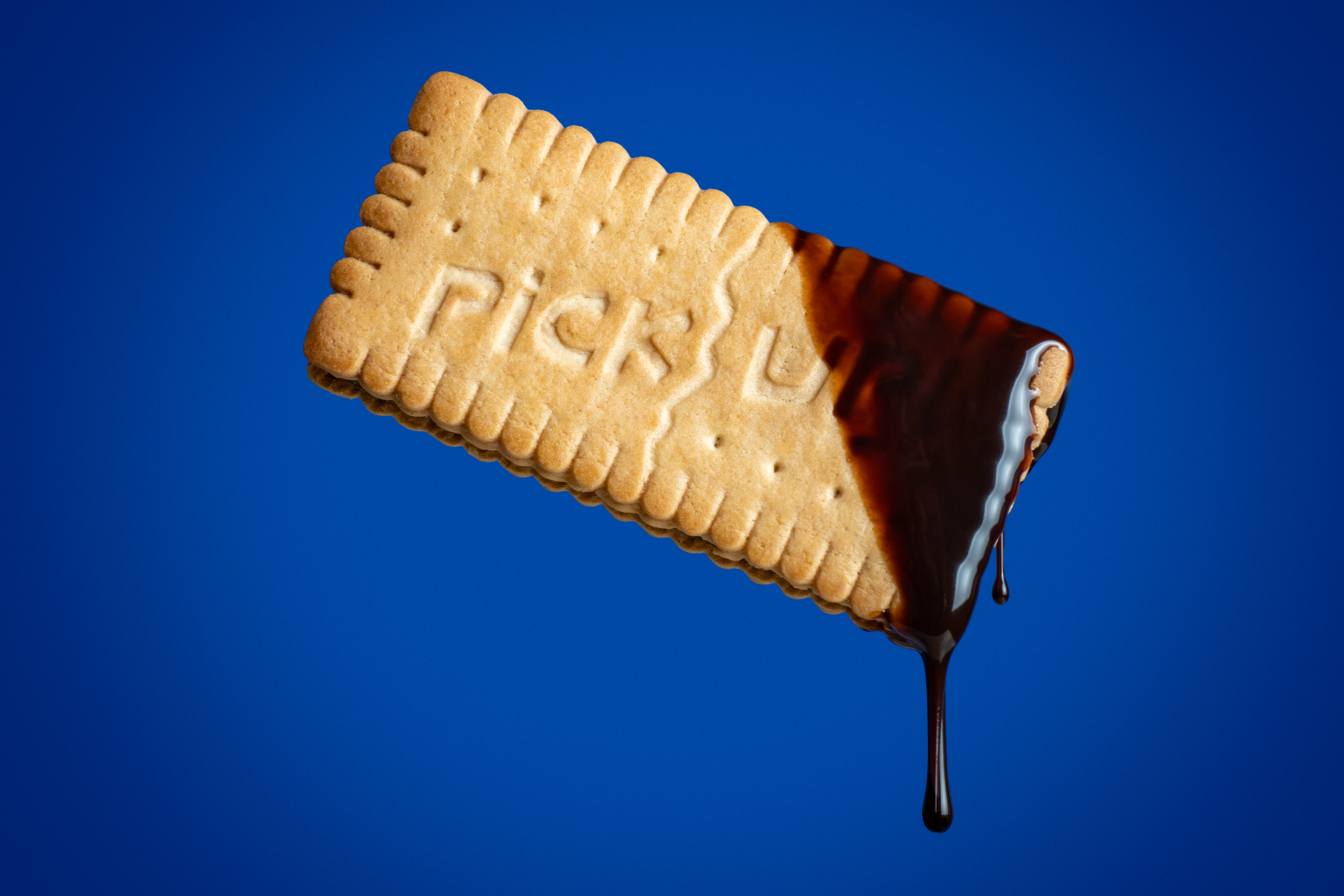 Pick Up biscuit dipped in chocolate with dripping chocolate on a bold blue background, food product photography by Photos by Lars.