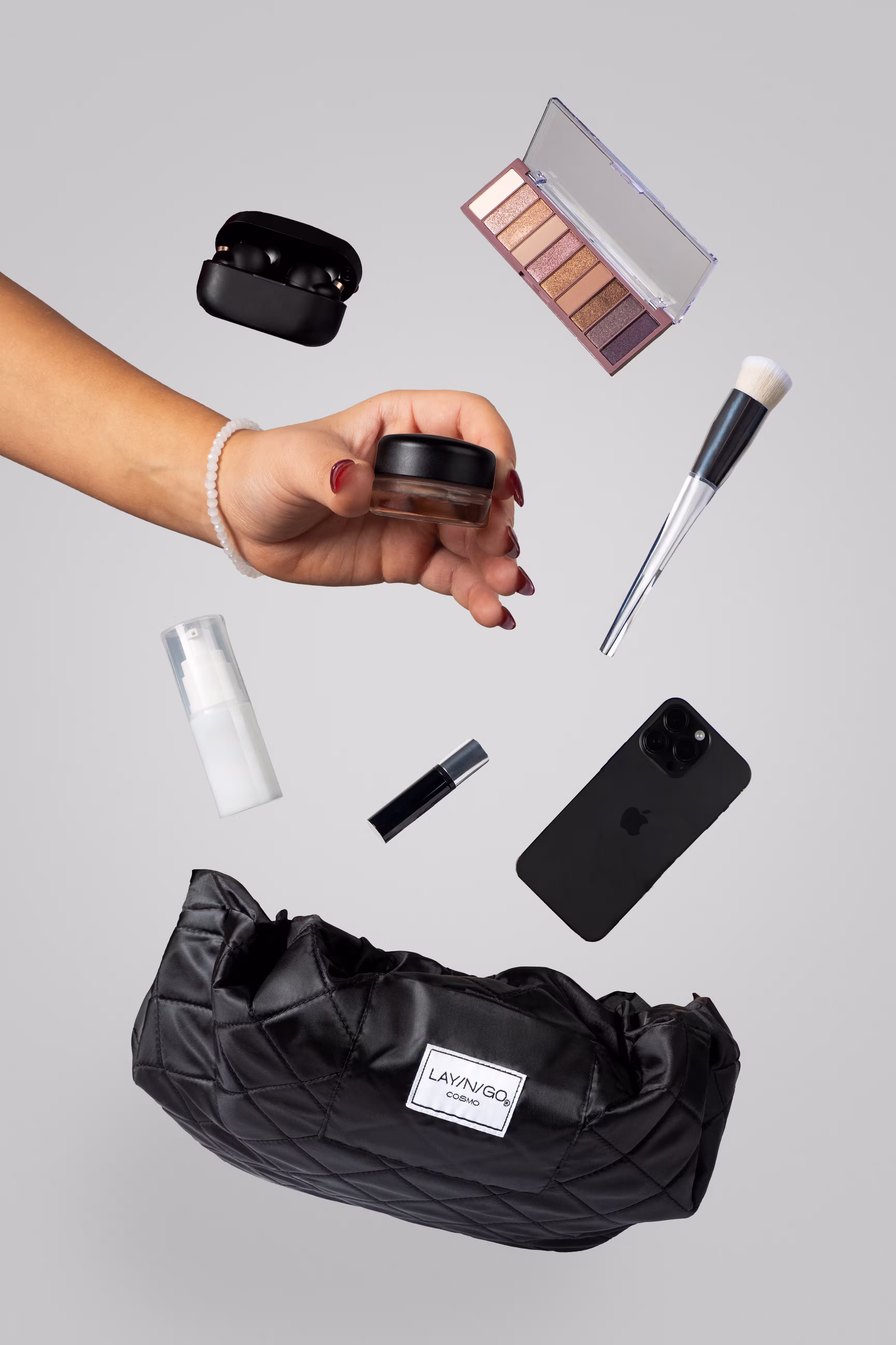 Hand holding small jar with makeup palette, brush, phone, and Lay-n-Go COSMO bag on gray, beauty product photography, Photos by Lars.