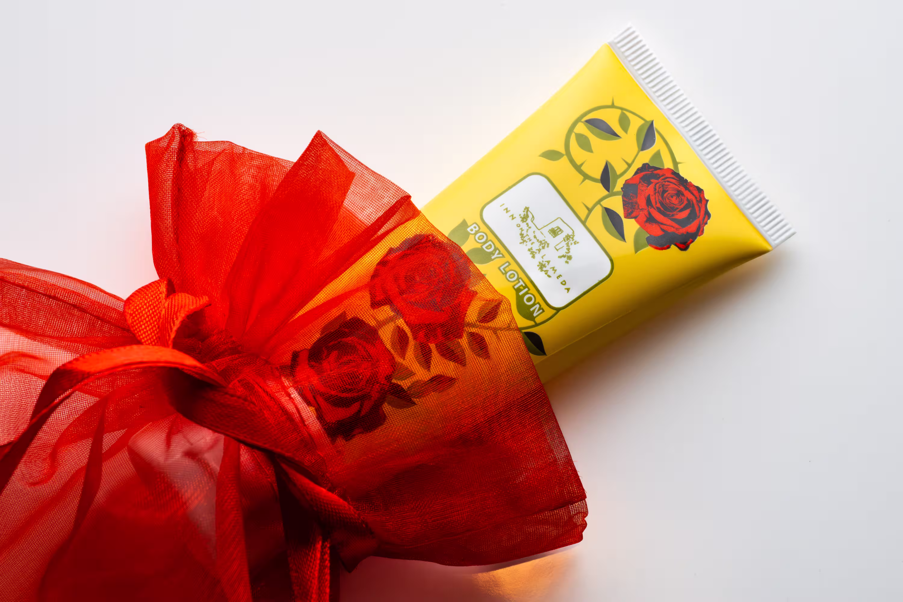 Yellow body lotion tube with red rose graphic next to red mesh gift wrap bow on white, product photography, Photos by Lars.