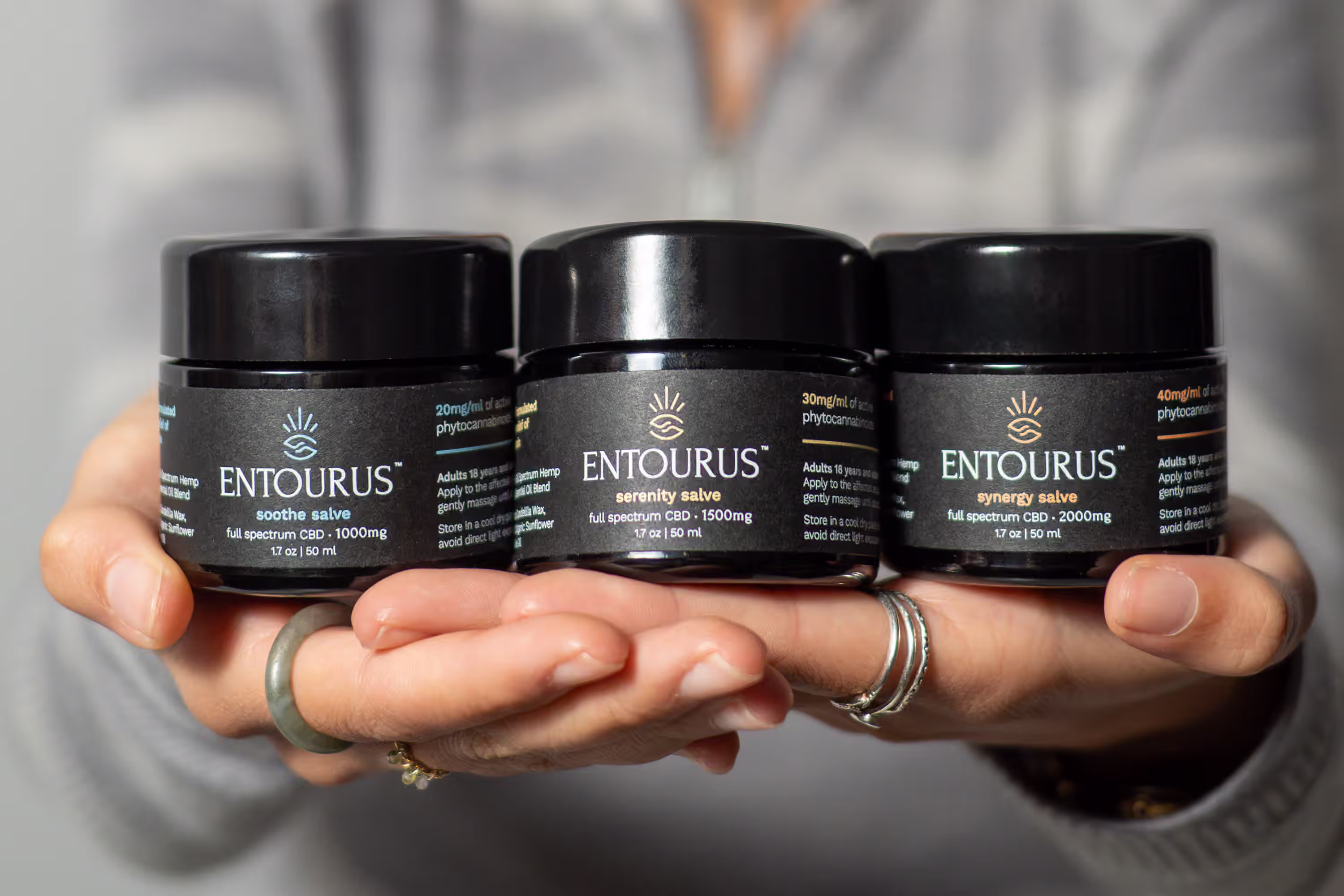 Three Entourus CBD salve jars held in two hands showing product scale and full product line for Amazon listing photography