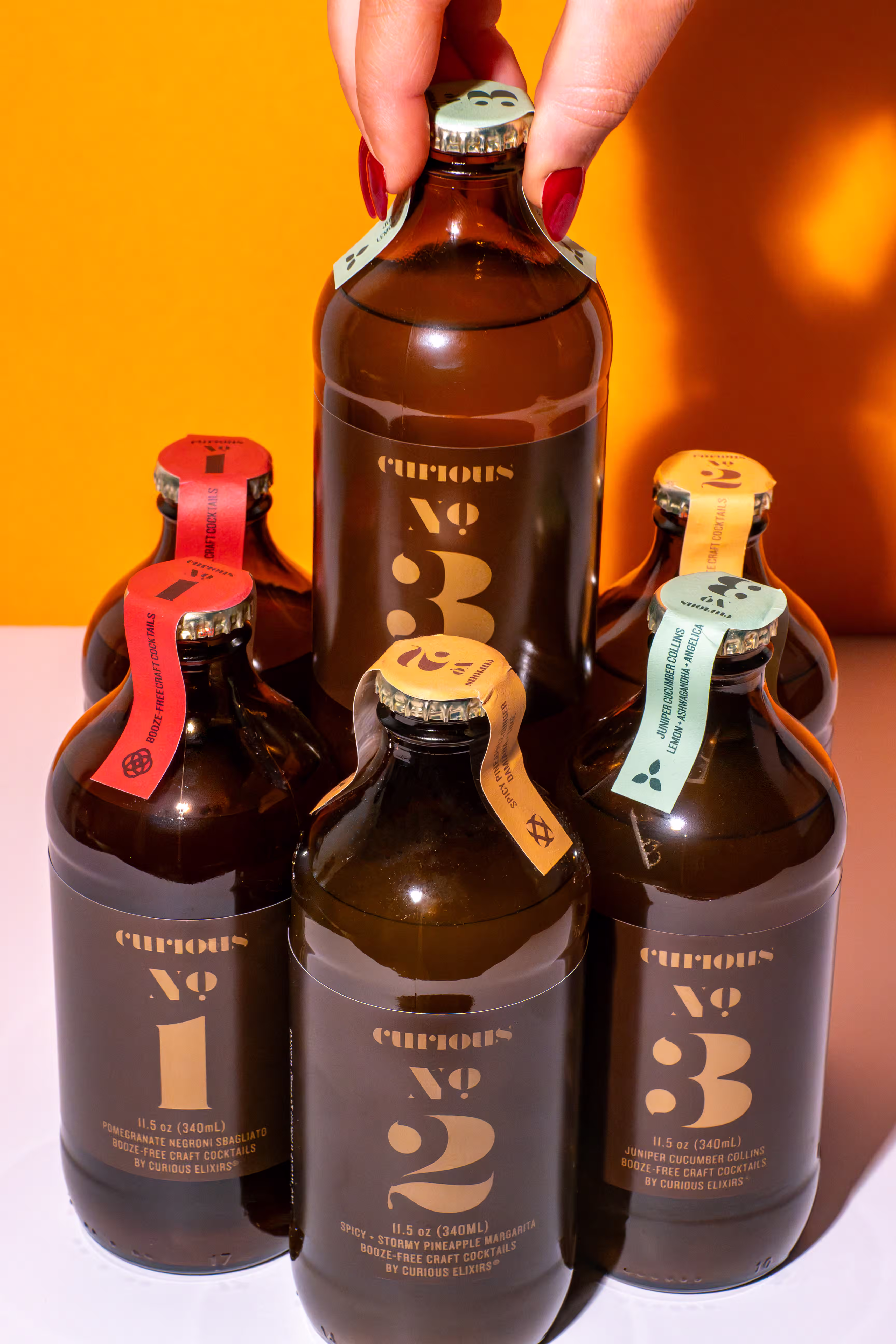 Hand reaching for a Curious Elixirs booze-free craft cocktail bottle among a styled collection of four flavors against an orange background