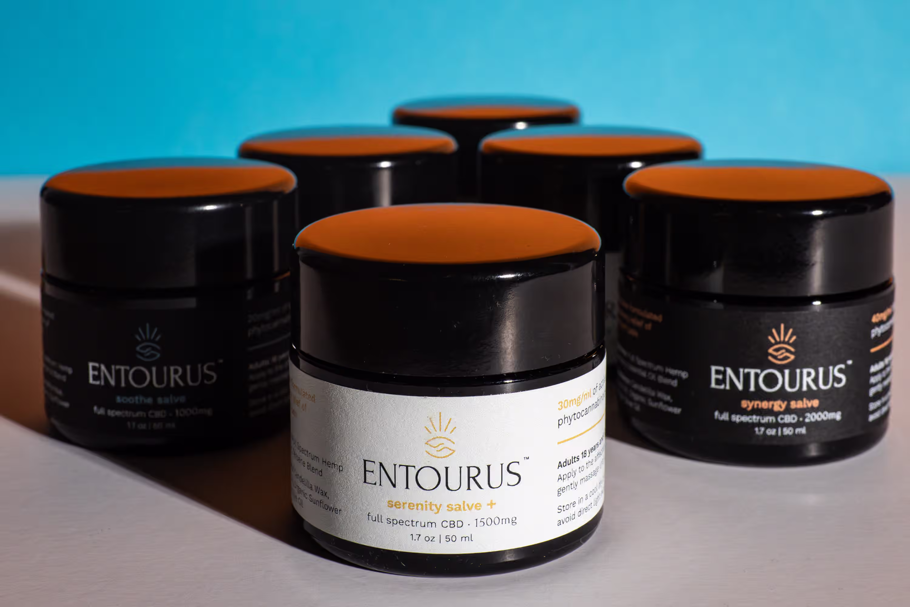 Six Entourus CBD salve jars photographed with consistent lighting and styling on a teal background showing product line consistency
