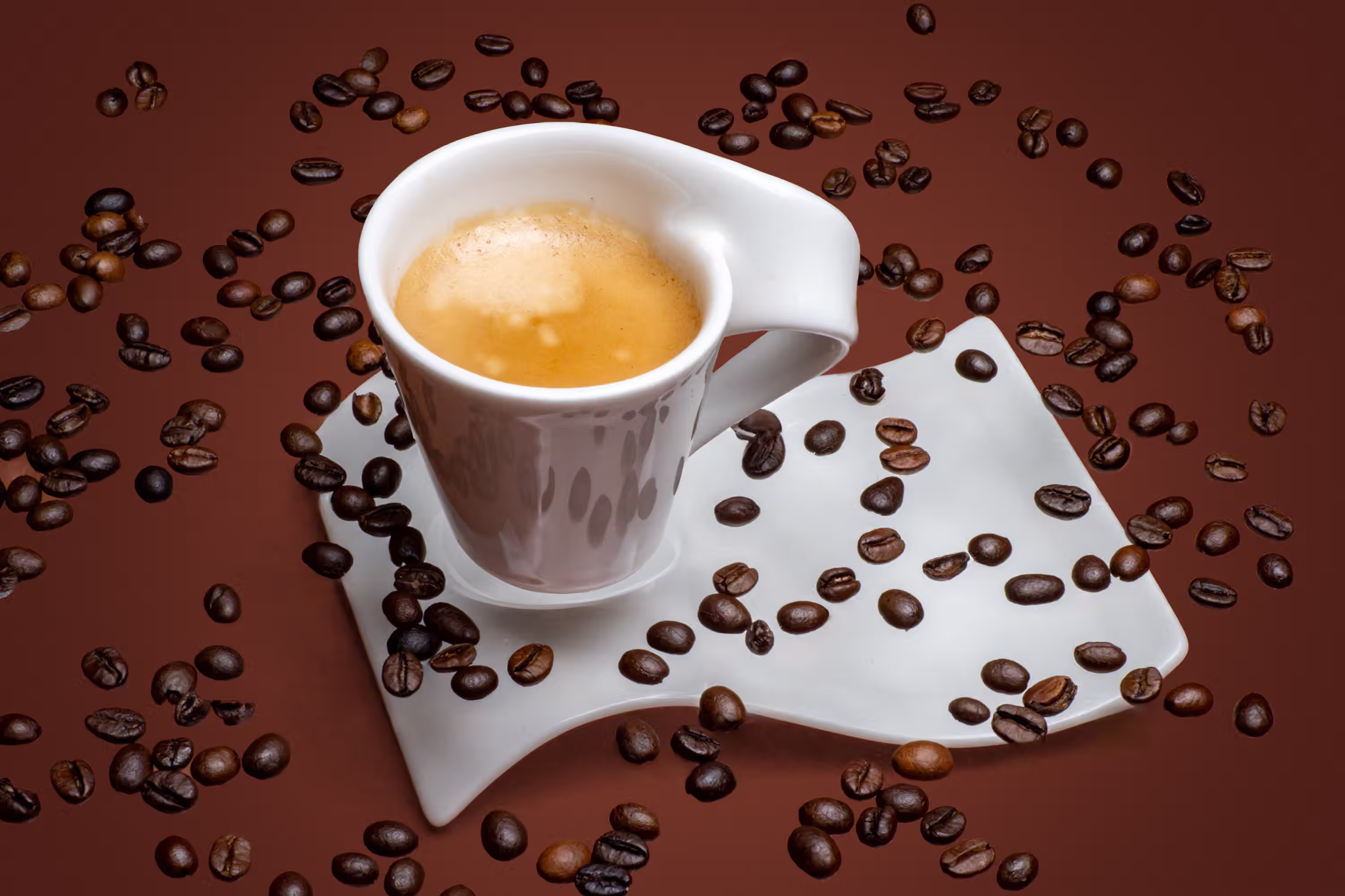 Espresso cup on a curved saucer surrounded by scattered coffee beans on a brown background in a styled product photograph