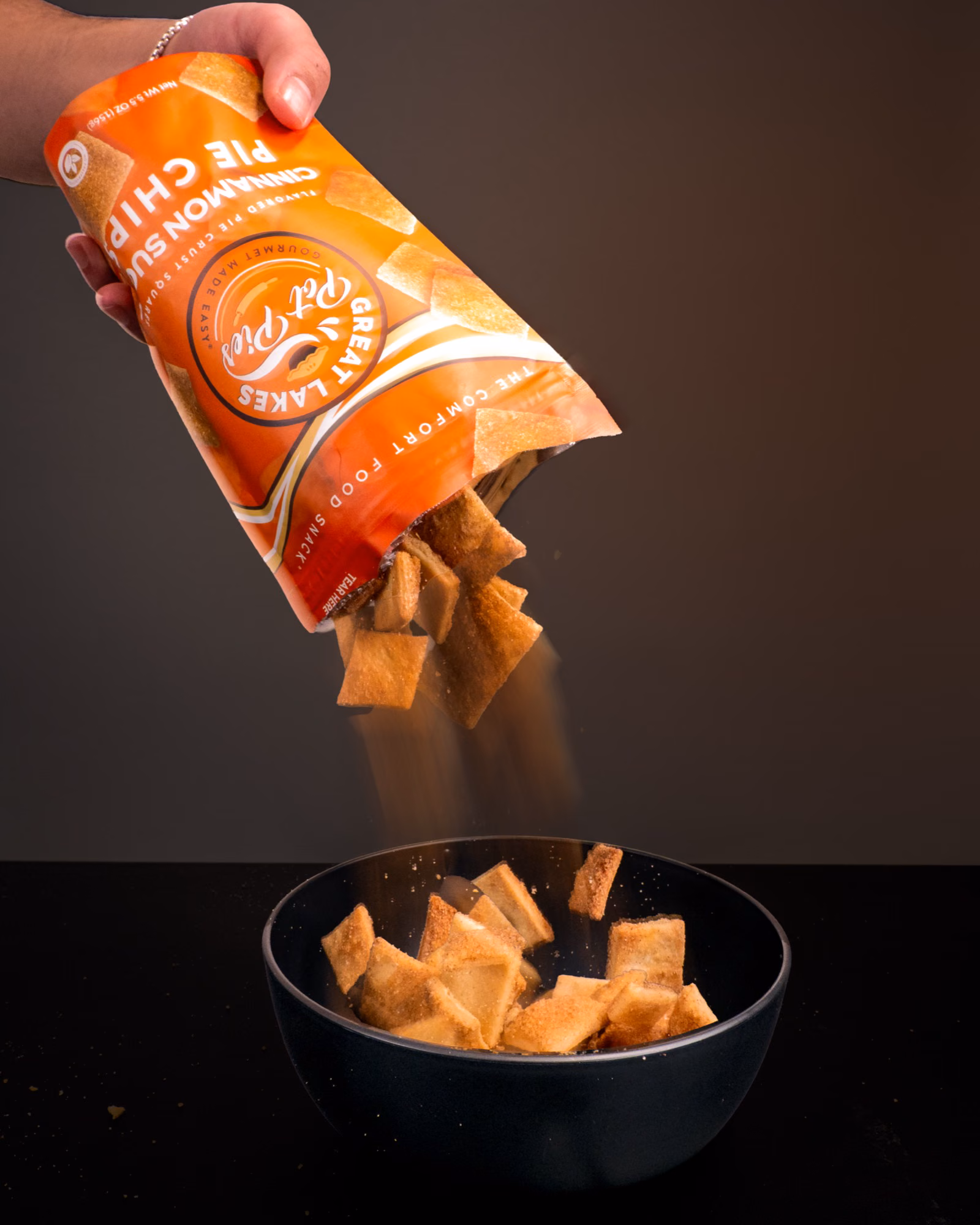 Great Lakes Pot Pies Cinnamon Sugar Pie Chips pouring from an orange bag into a black bowl in a studio motion photo.