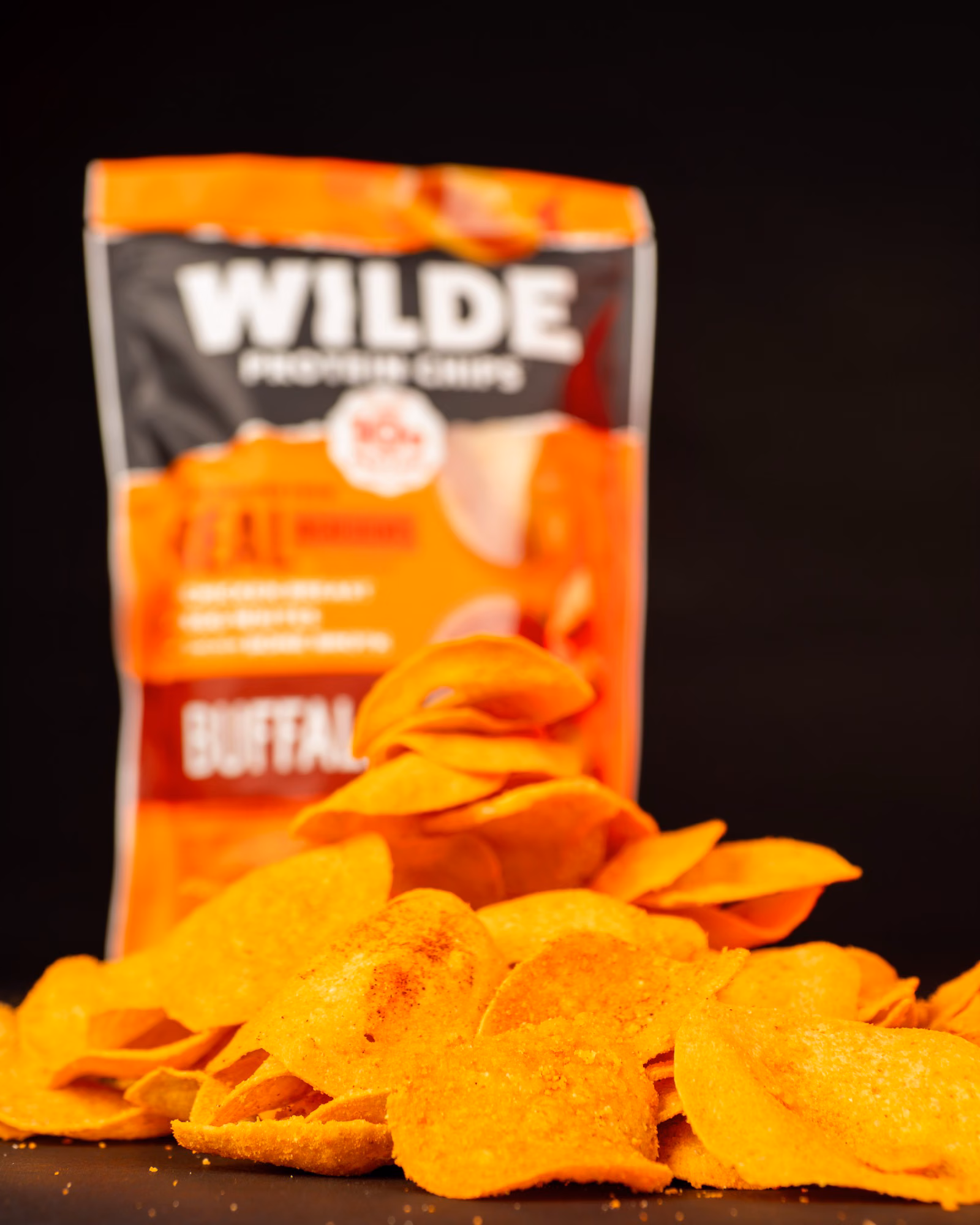 Close-up of orange Wilde Protein Chips piled in front of a blurred Wilde Buffalo protein chips bag.