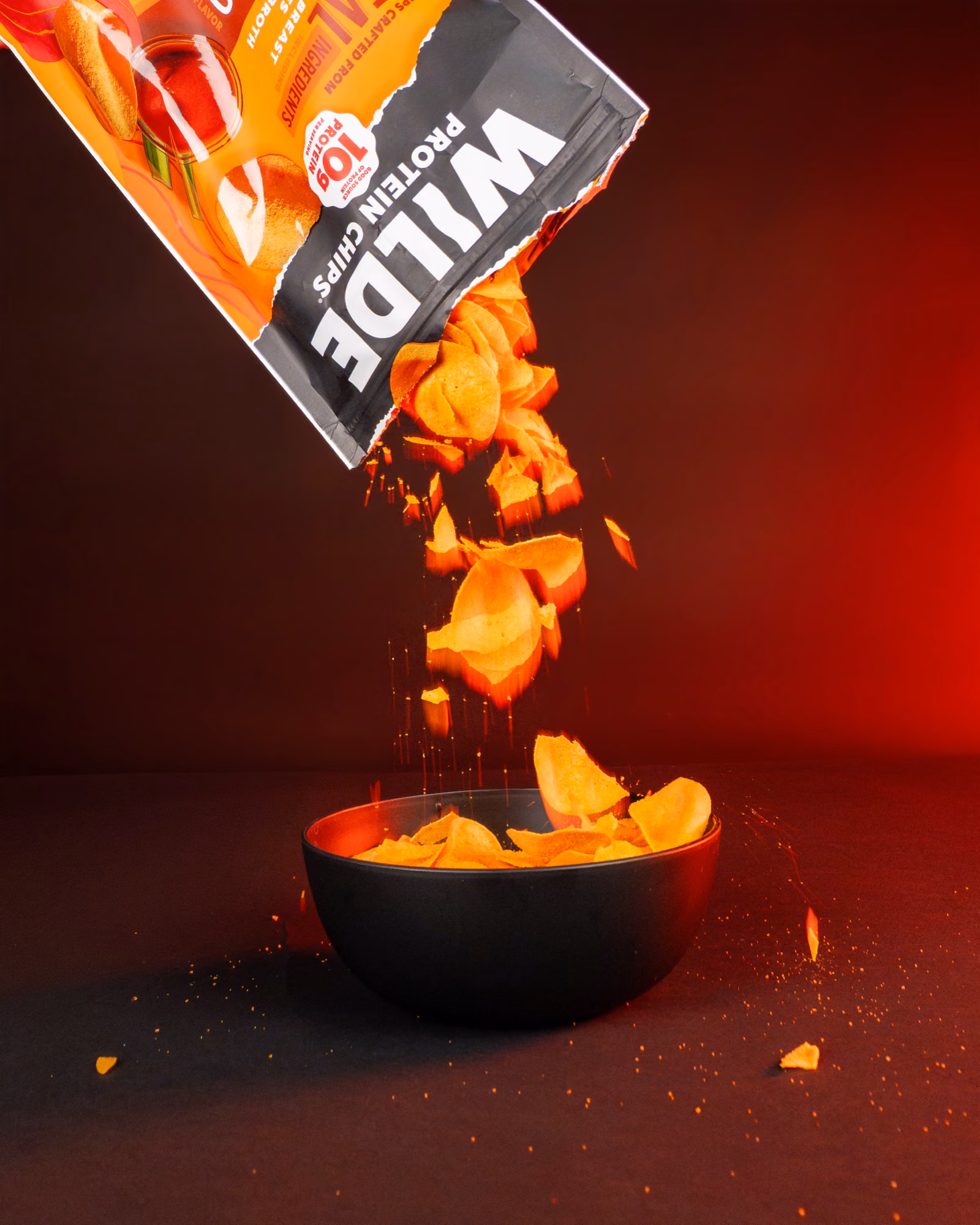 Wilde Protein Chips pouring from an orange bag into a black bowl with dramatic red studio lighting.